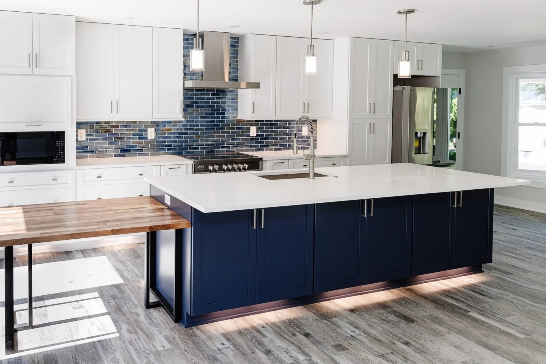 Modern kitchen with white cabinets, a navy blue island with a white countertop, a wooden side table, stainless steel appliances, blue tiled backsplash, pendant lighting, and large windows.