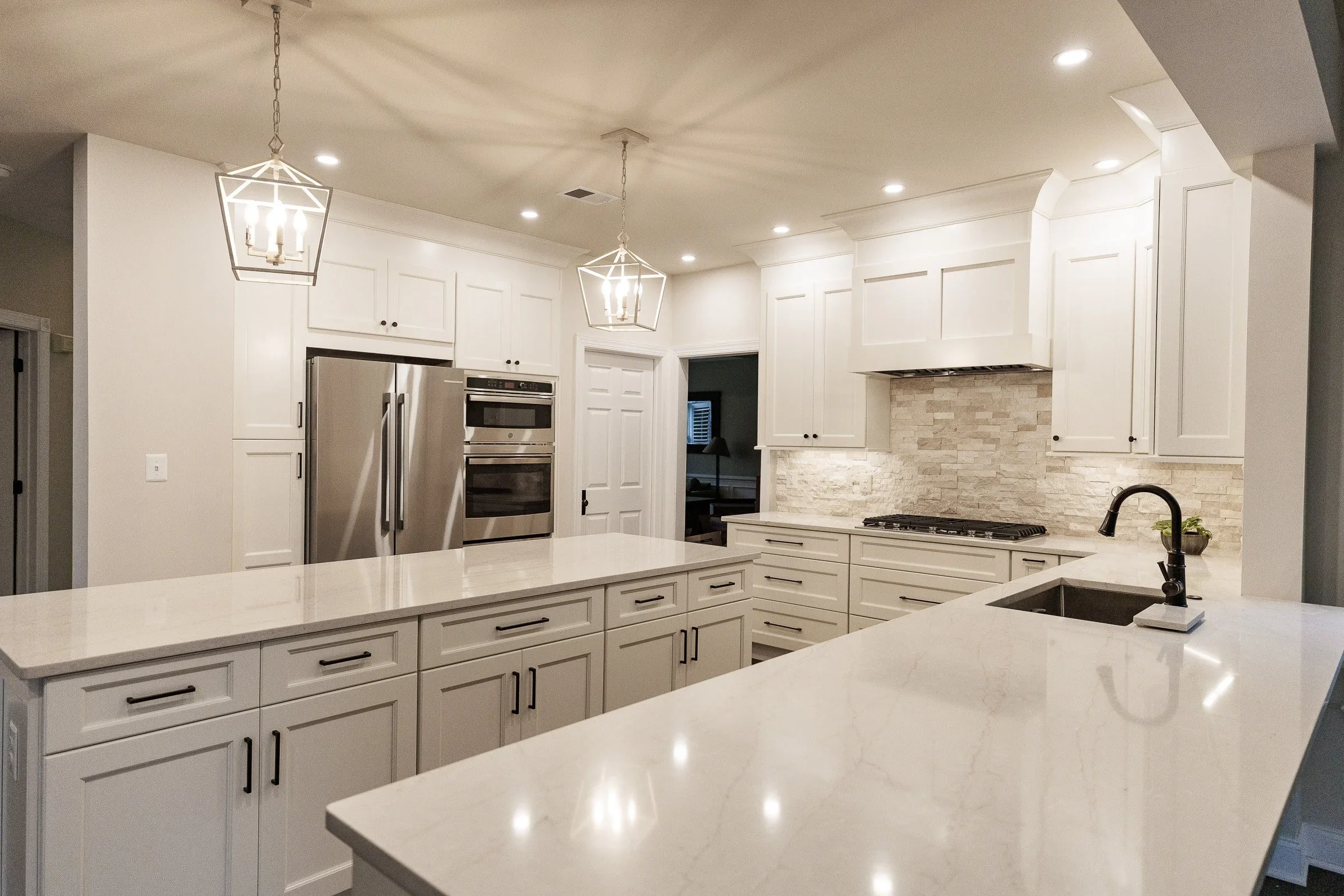 Modern kitchen with white cabinets, stone backsplash, stainless steel appliances, and a large white island with a sink. Two geometric pendant lights hang from the ceiling, and recessed lighting is also present.