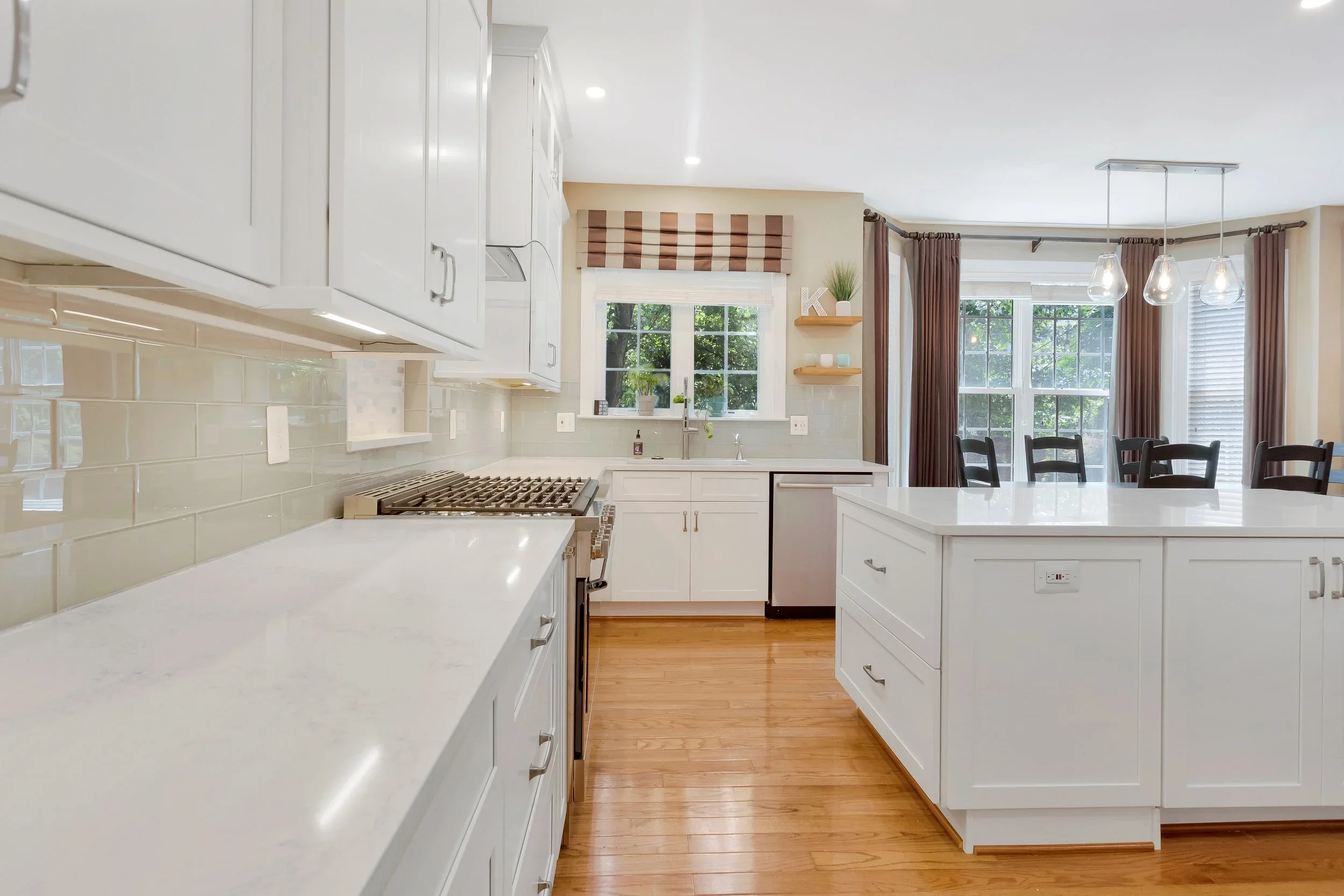 A bright, modern kitchen with white cabinets, a beige subway tile backsplash, and hardwood floors. Large windows with brown curtains and a striped window treatment let in natural light. There are pendant lights hanging over a white island and a dinin