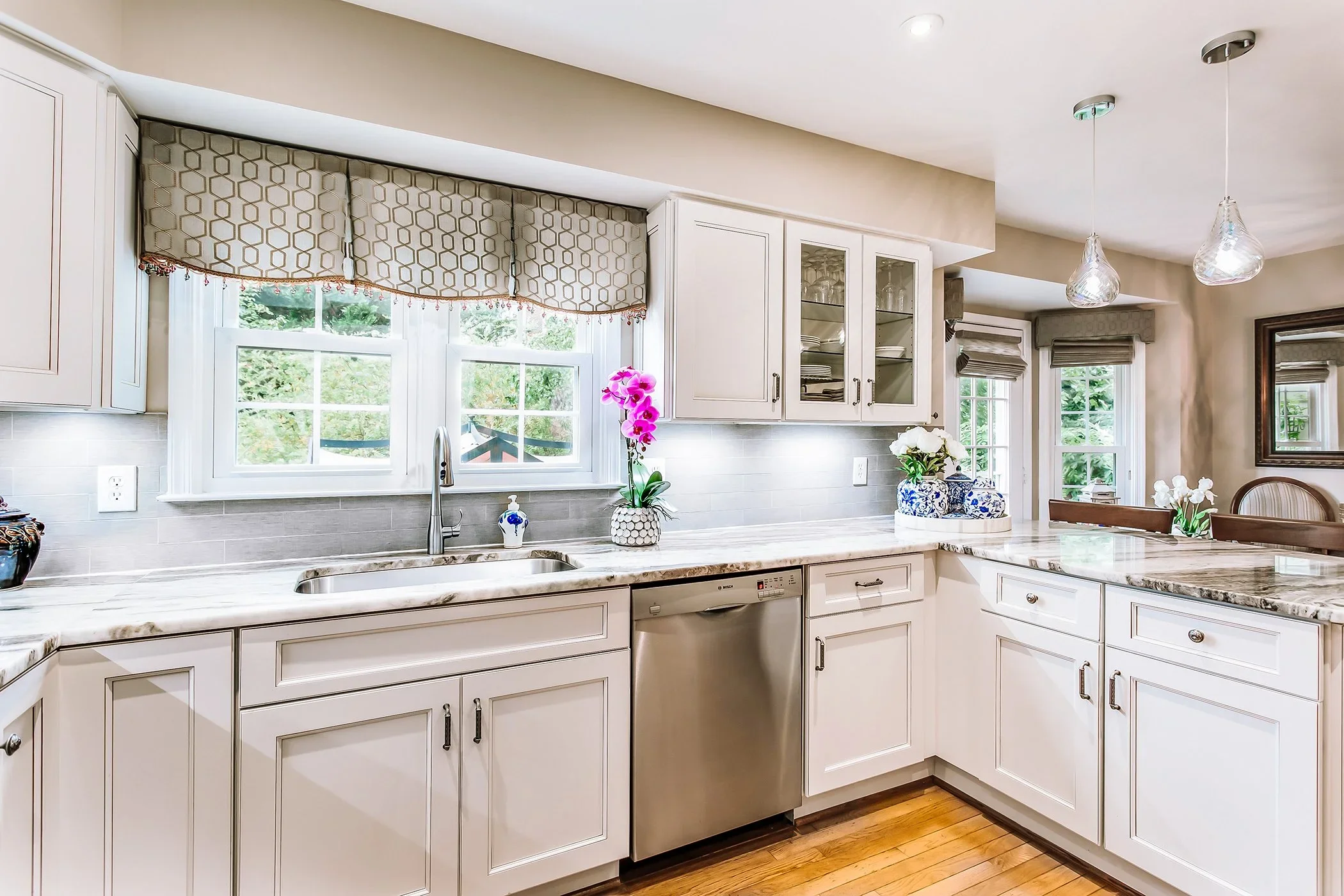 A bright kitchen with white cabinets, a marble countertop, and a window with hexagonal-patterned valance. There are pink and white flowers in vases, and hanging glass pendant lights.