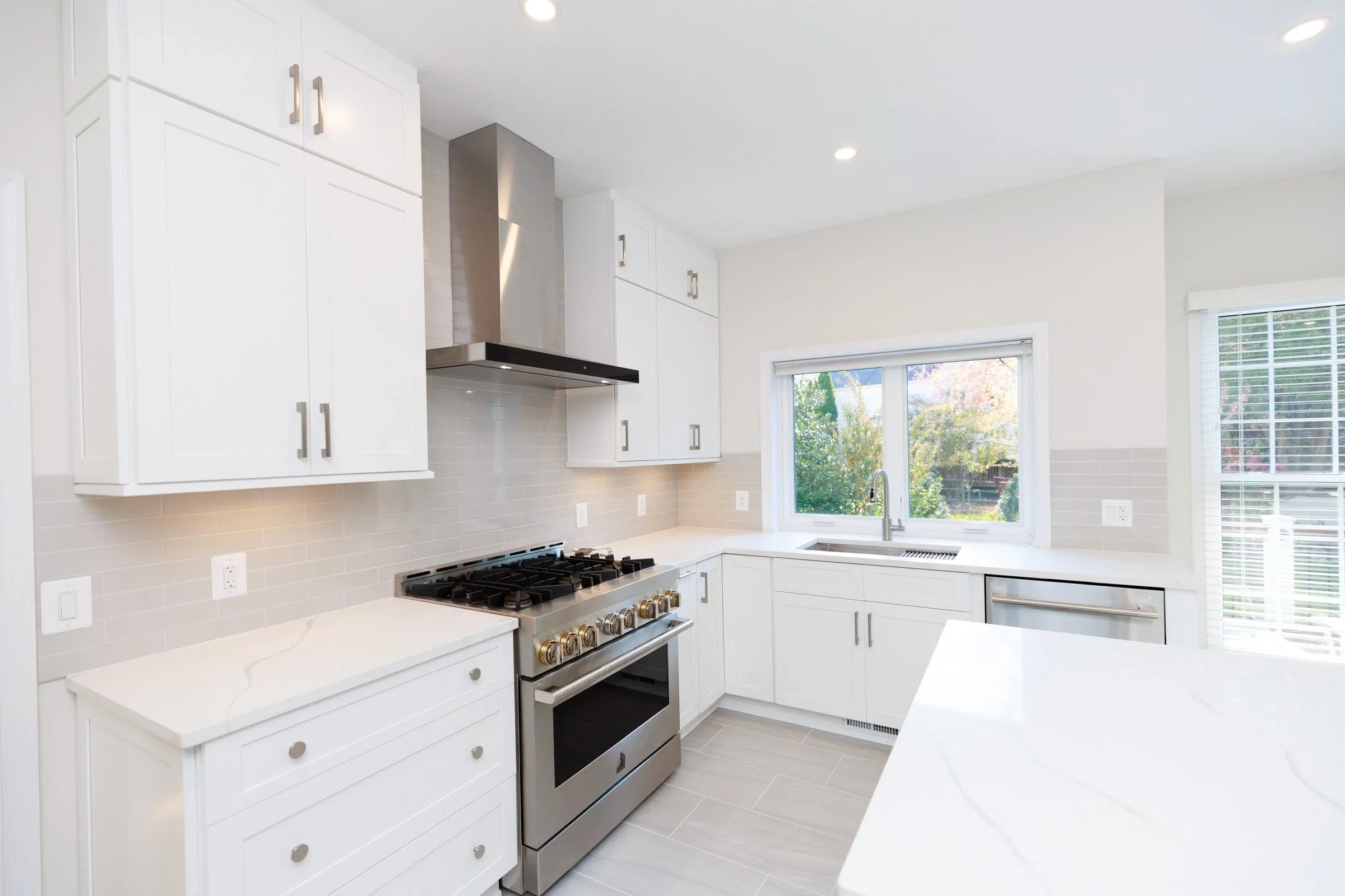 Modern white kitchen with stainless steel stove, range hood, and large window with a view of trees outside.