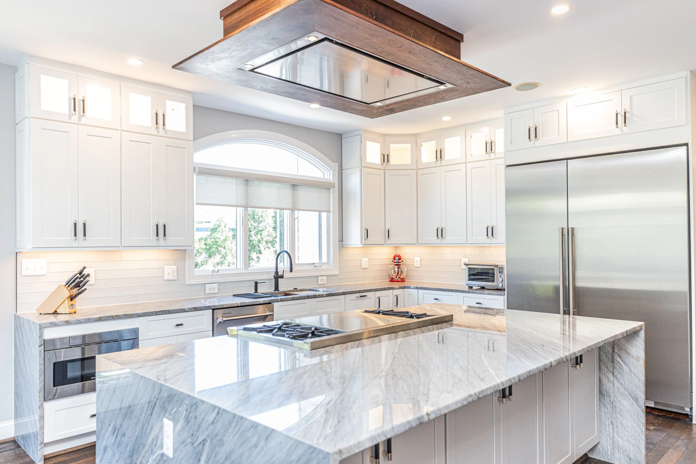 Modern kitchen with white cabinets, marble countertop island, stainless steel refrigerator, and a window with blinds.