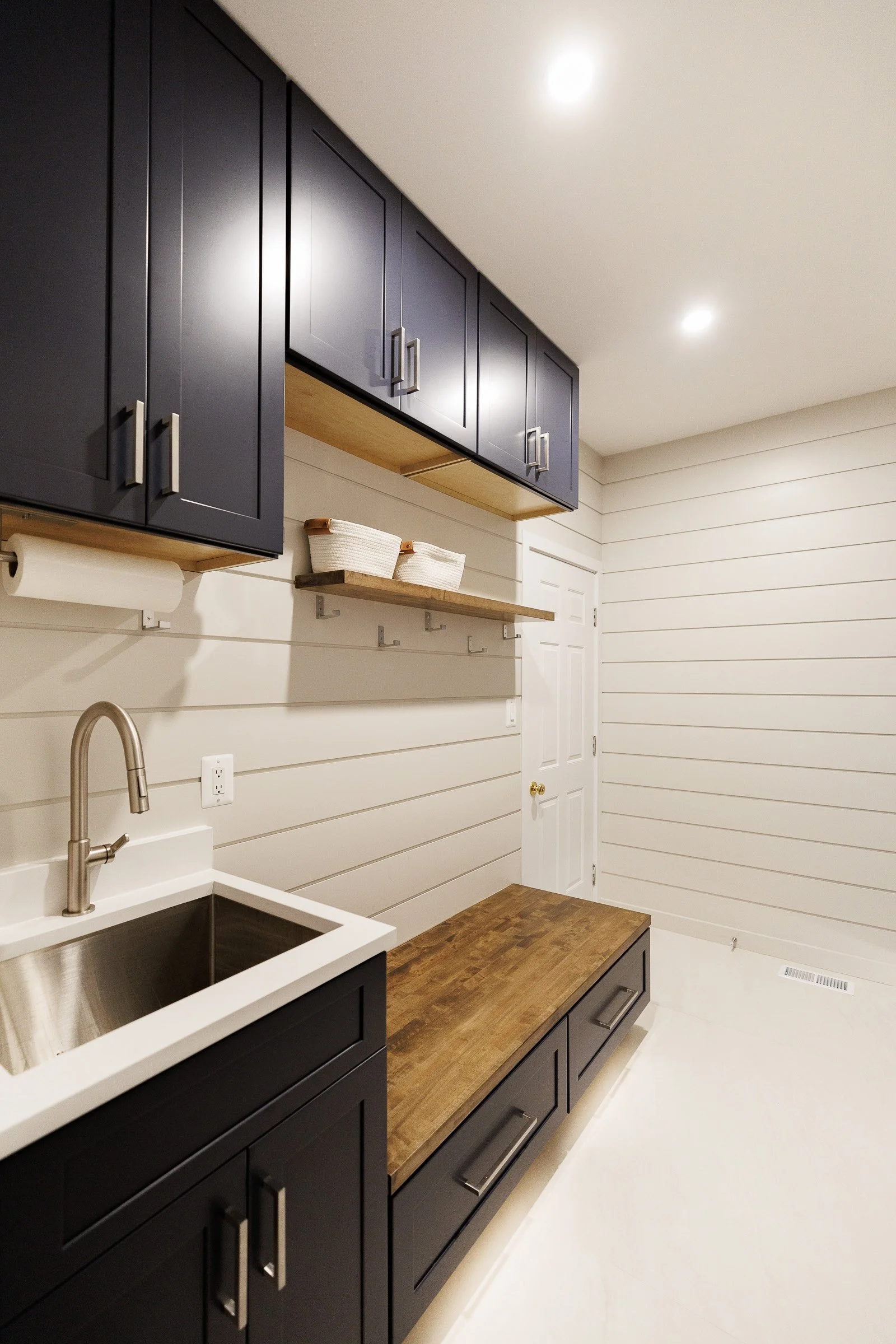 Interior of a modern kitchen with white shiplap walls, dark navy cabinets with stainless steel handles, a wooden countertop, and a small open shelf with baskets. There is a white farmhouse sink with a stainless steel faucet and a paper towel roll hol