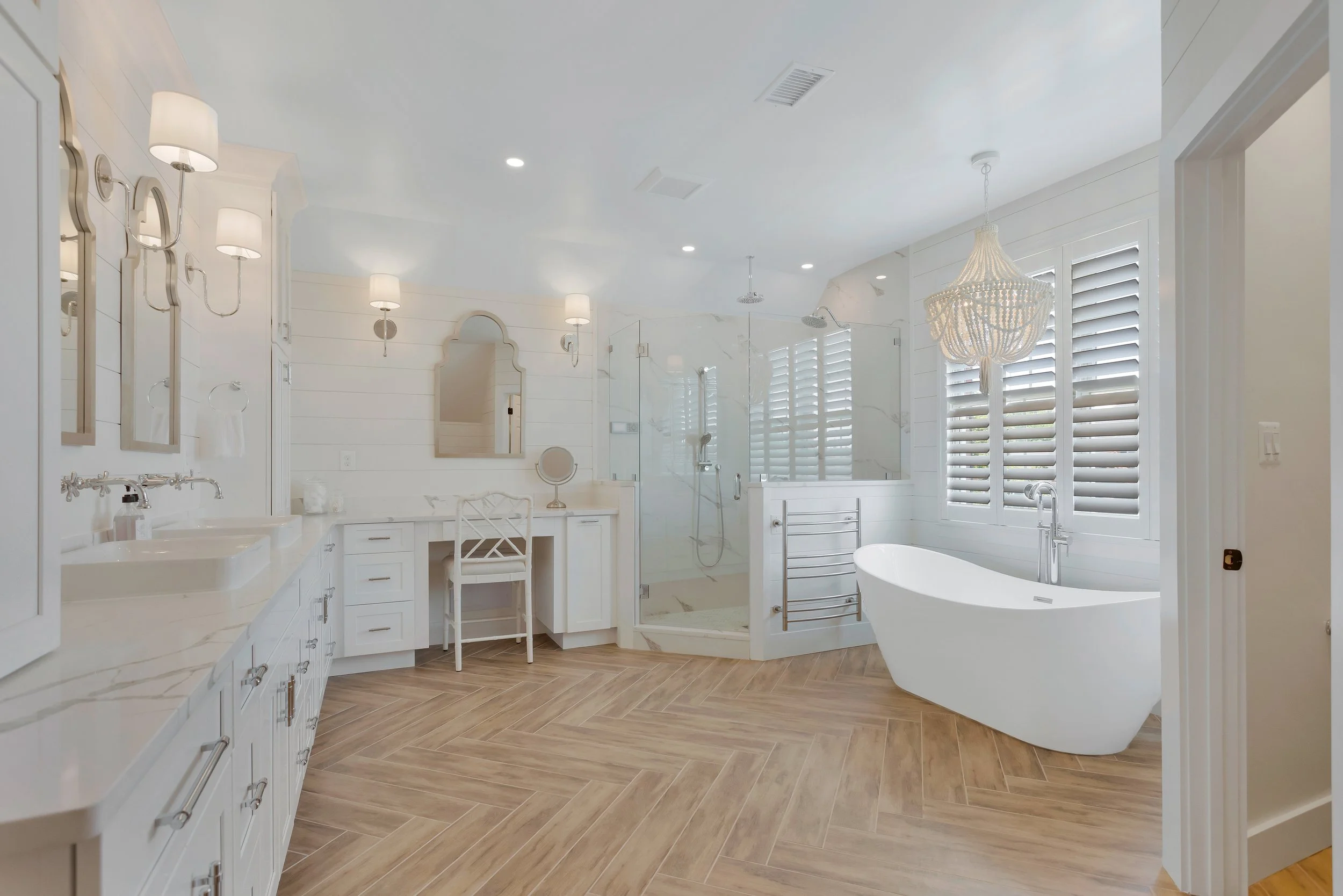 A modern, white bathroom with a large bathtub, a walk-in shower with glass door, white cabinetry, and a chandelier. Louvered shutters cover the windows.