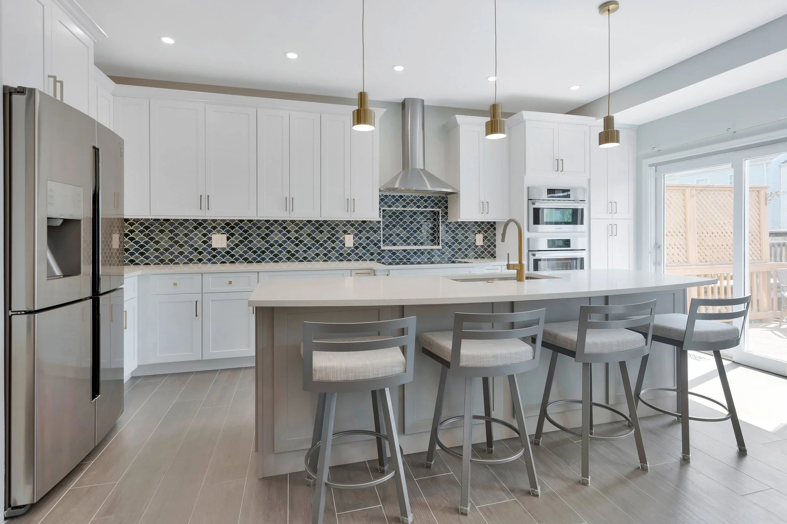 Modern kitchen with white cabinets, dark patterned backsplash, stainless steel refrigerator, built-in oven, large island with seating, pendant lighting, sliding glass door to patio.
