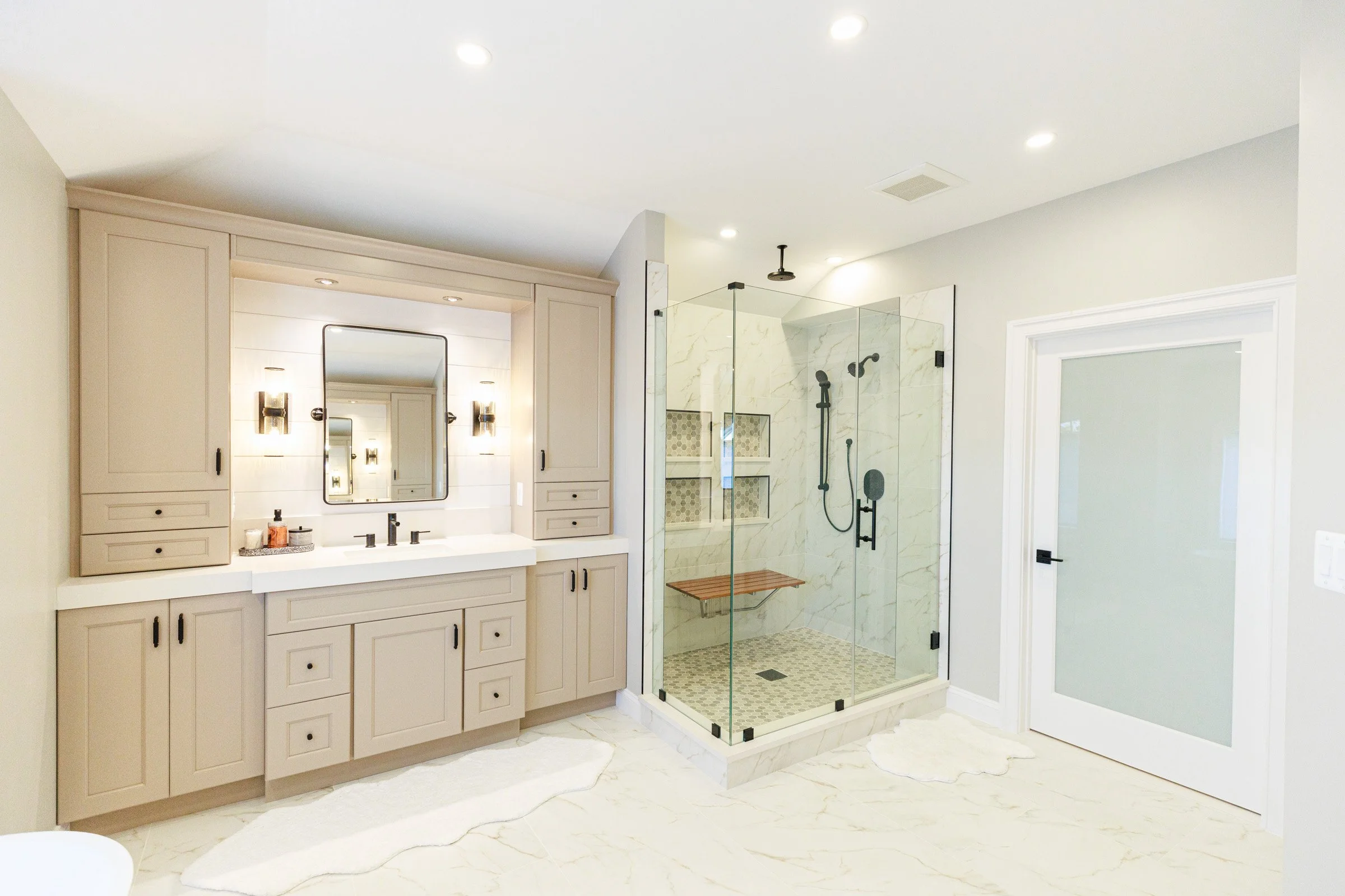 A modern bathroom with a beige vanity, a large mirror, black fixtures, and a walk-in shower enclosed by glass with marble tiles and a wooden bench inside.