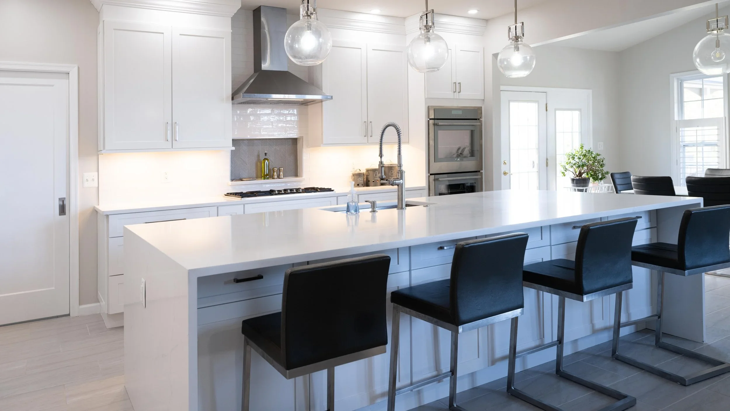 Modern white kitchen with large island, black barstools, stainless steel appliances, pendant lighting, and a door leading outside.