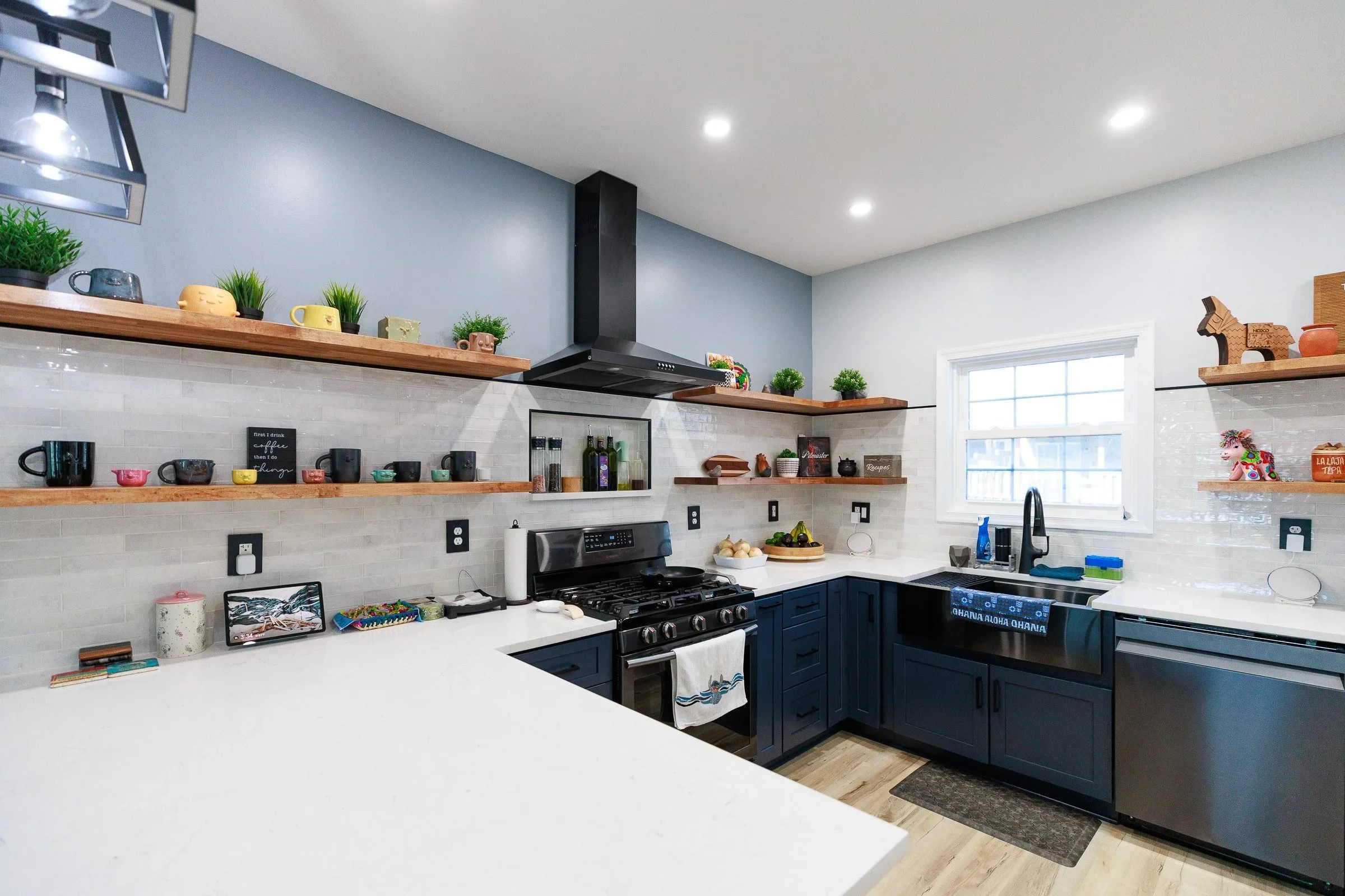 A modern kitchen with white countertops, blue cabinets, and a white tiled backsplash. Open wooden shelves hold potted plants, cups, and decorative items. There are black electrical outlets, a stove, a sink, and various kitchen essentials on the count