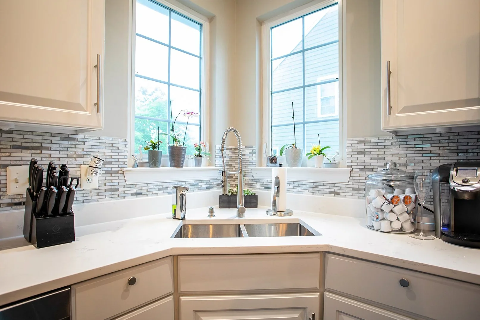 Kitchen sink area with two large windows, plants on the windowsill, a black knife block, and a container of coffee pods.