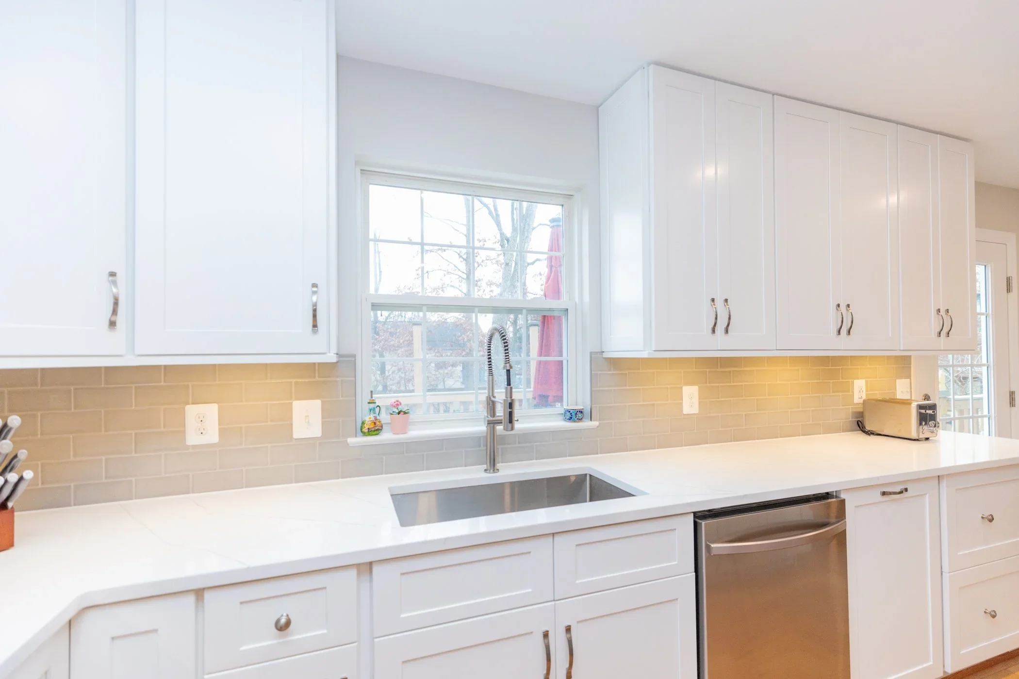White kitchen cabinets with silver handles, beige subway tile backsplash, white countertop, window above the sink with a tree outside, small potted plant, soap, and a toaster on the counter.