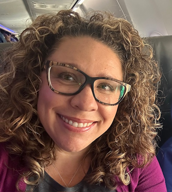 A woman with curly hair and glasses smiling on an airplane.