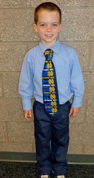 Young boy wearing a light blue dress shirt, University of Michigan tie with yellow and blue patterns, navy pants, and black shoes standing against a gray brick wall.