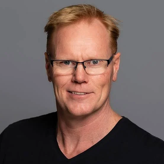 A headshot of a middle-aged man with red hair, wearing glasses and a black shirt, against a gray background.