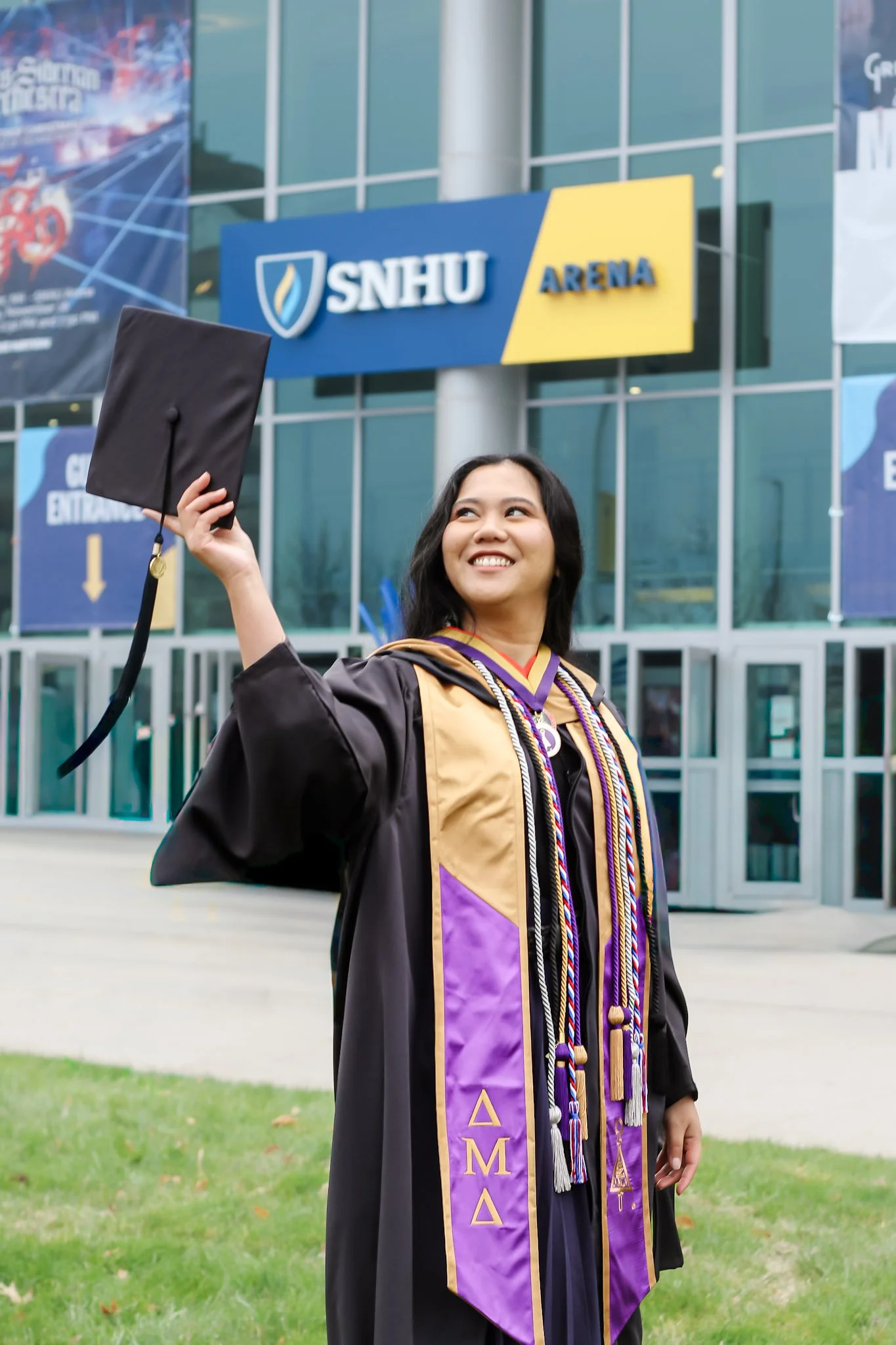 A young woman in graduation cap and gown holding her cap in the air outside of SNHU Arena, celebrating her graduation.