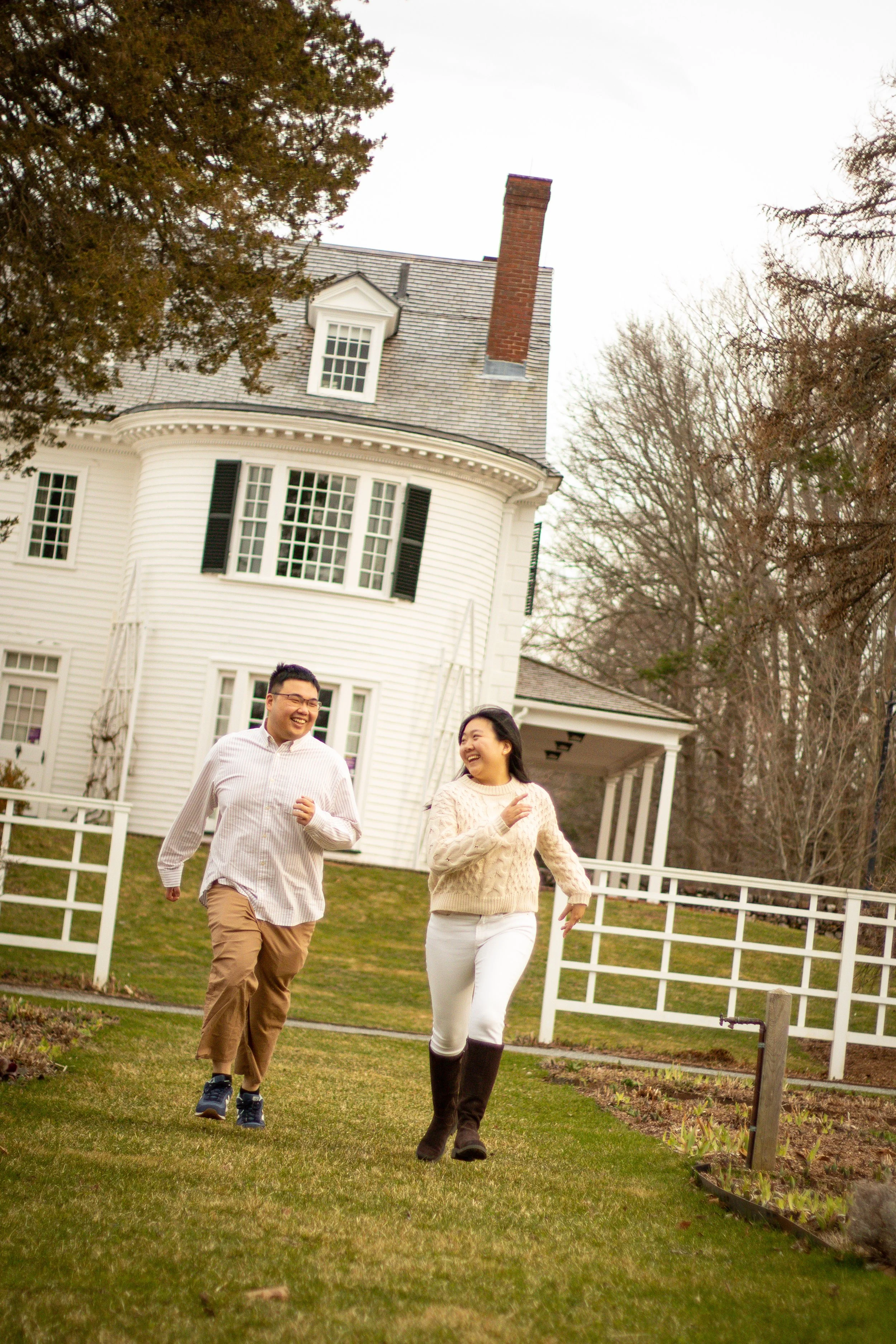 A man and woman running and smiling in front of a large white house with black shutters and a manicured lawn.