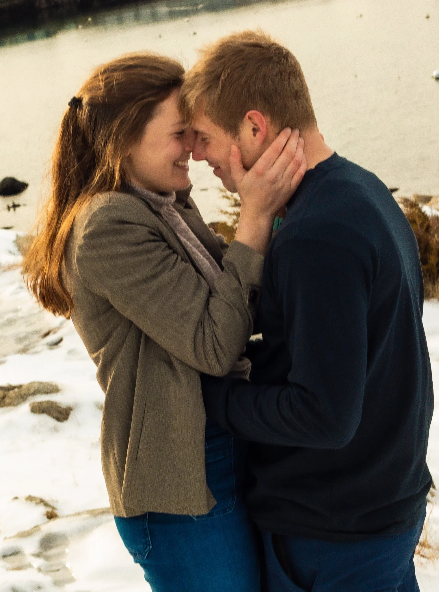 A couple standing close together outdoors on a snowy ground, touching foreheads, with the woman holding the man's face gently, smiling.