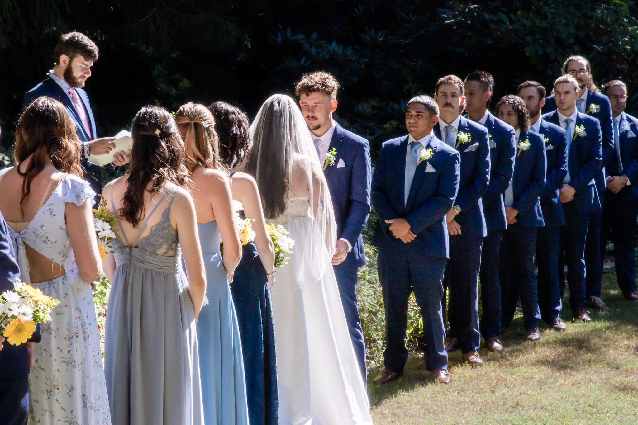 A wedding ceremony outdoors with the bride and groom facing each other, surrounded by bridesmaids and groomsmen.