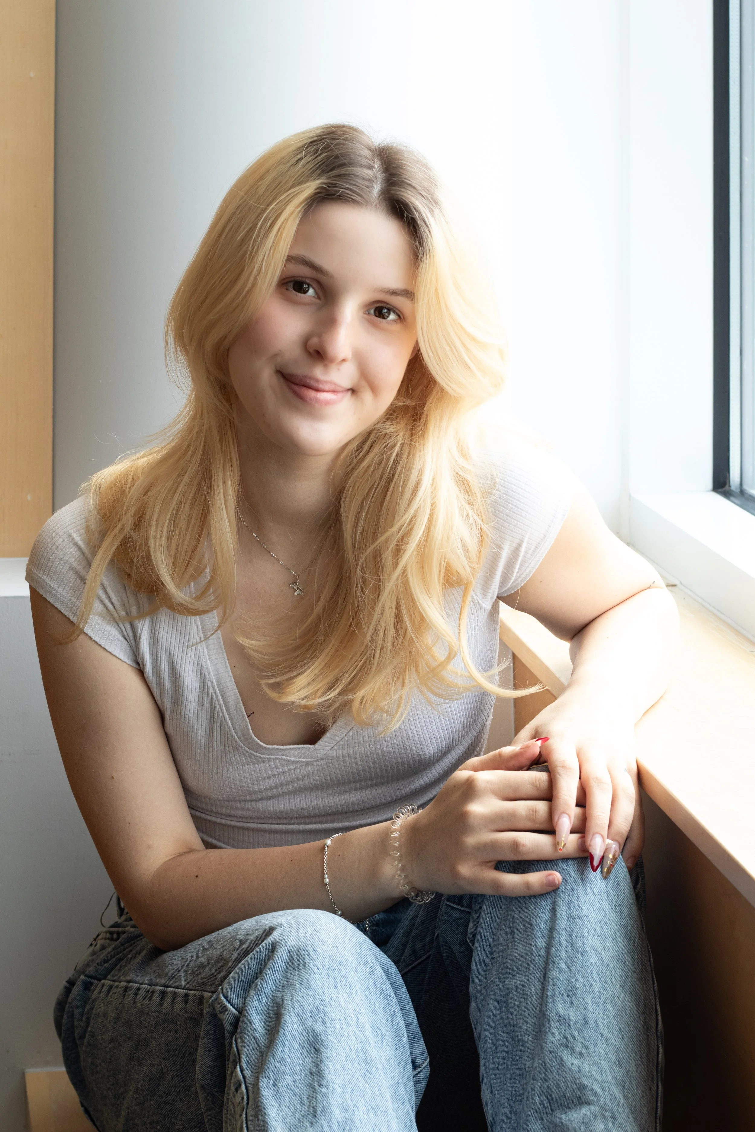 Senior Portraits, Wenham, MA. A young woman with long blonde hair sitting by a window, smiling slightly, holding her hands together on her lap, wearing a beige t-shirt and light blue jeans.