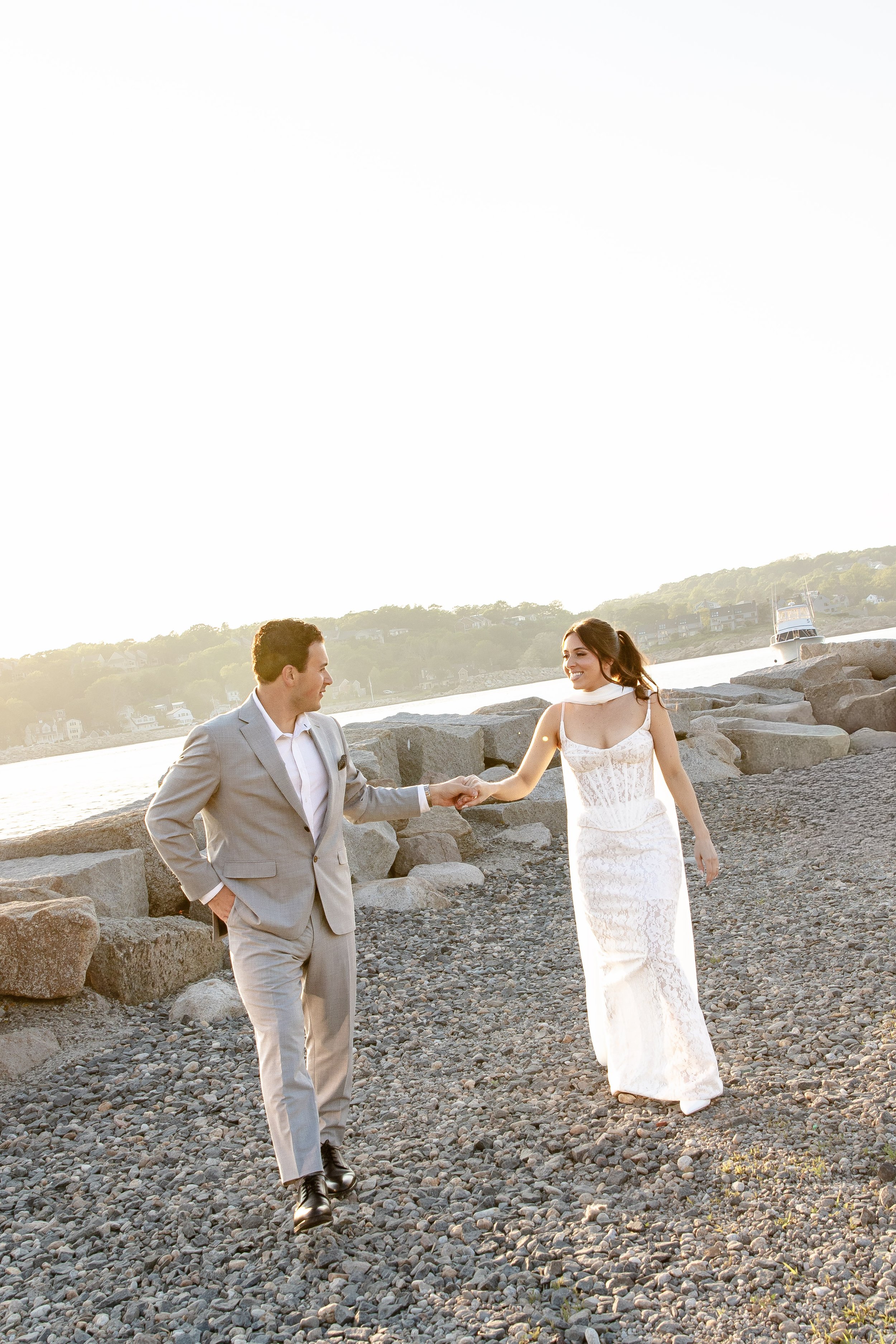 A man in a gray suit and a woman in a white lace dress are holding hands and smiling while walking on a rocky beach with boats and houses on a hillside in the background during sunset.