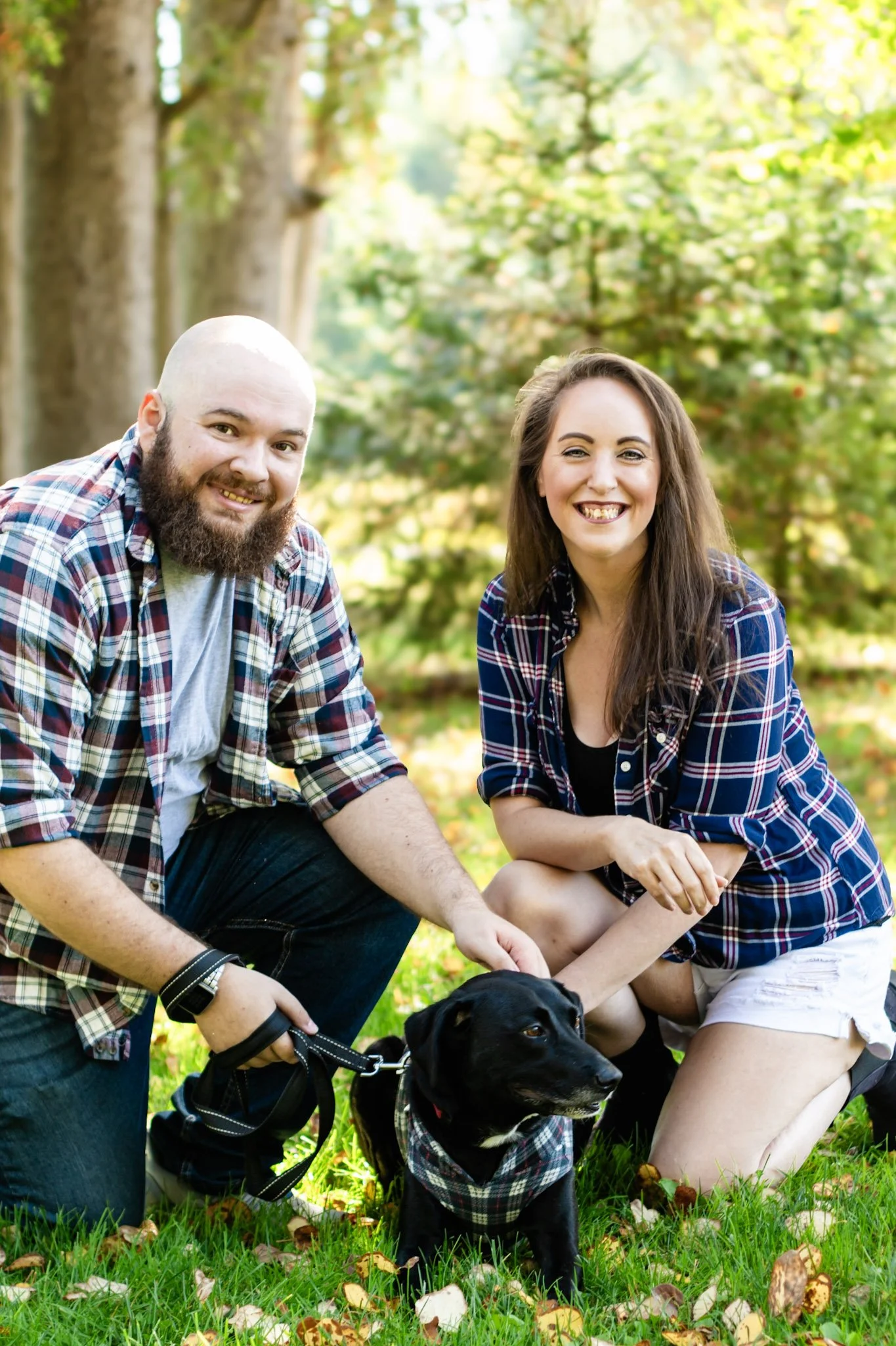 A man and woman with a black dog sitting on grass in a park, surrounded by trees with fall leaves.