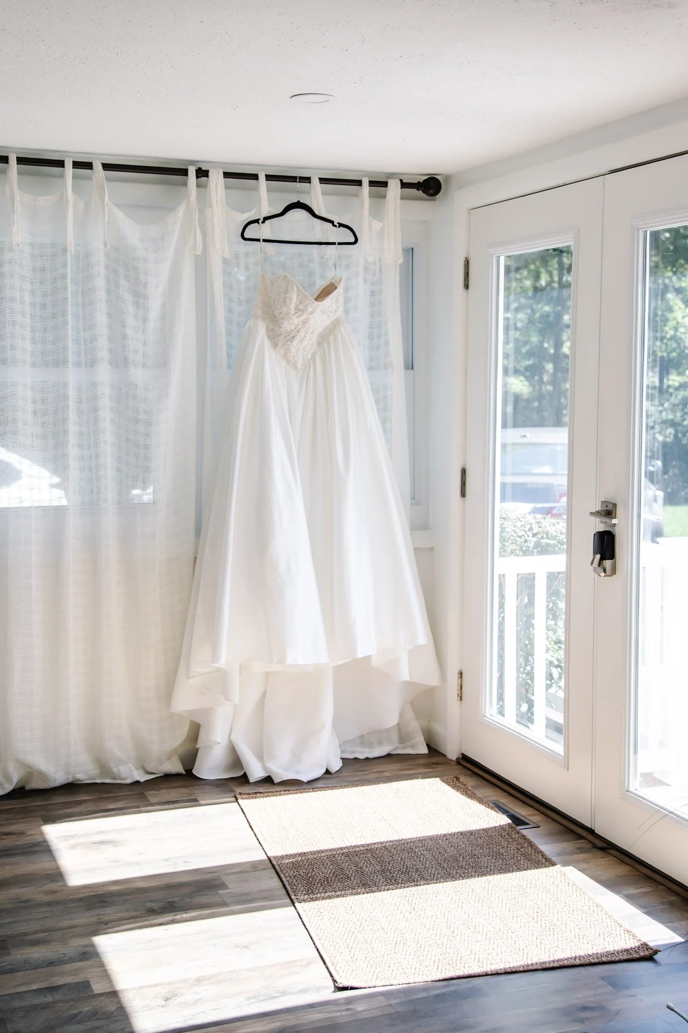 A white wedding dress hanging on a black hanger inside a bright room near a glass door with sunlight streaming in.