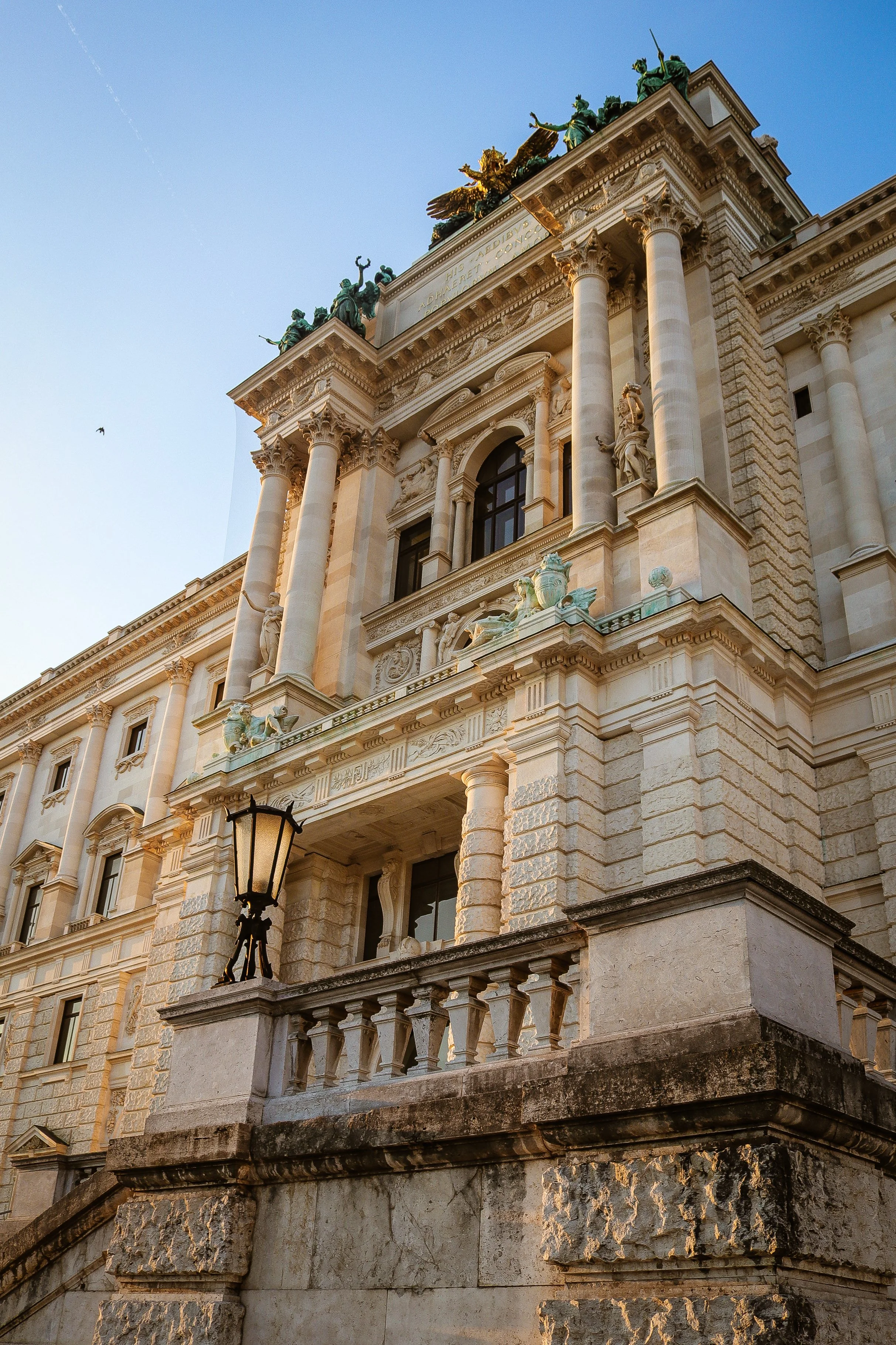 A historic building with classical architecture, featuring ornate columns, detailed sculptures, and a dramatic rooftop statue of a chariot pulled by winged horses, bathed in warm sunlight.