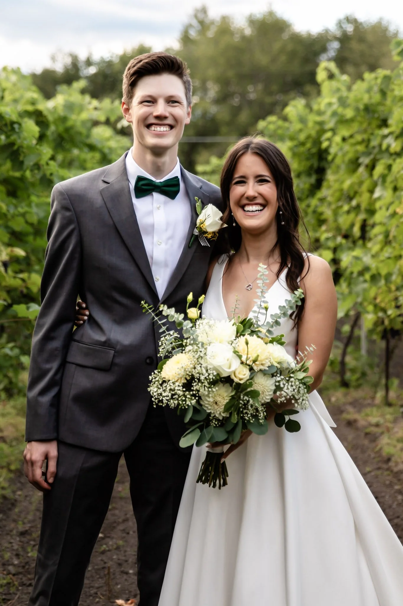 A happy bride and groom smiling outdoors in a vineyard, with the bride holding a bouquet of white and yellow flowers.