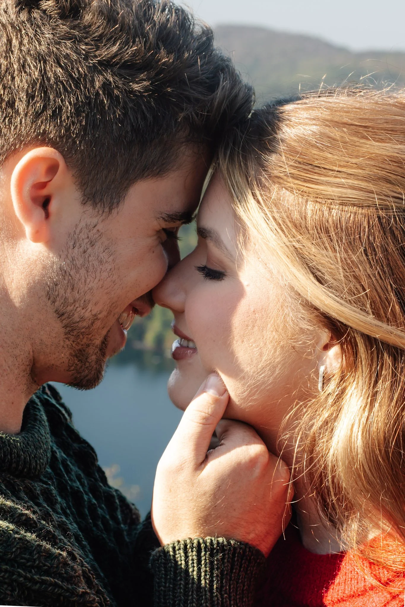 A man and woman with their foreheads and noses touching, smiling, with the man's hand gently holding the woman's chin, outdoors near a body of water and trees in the background.