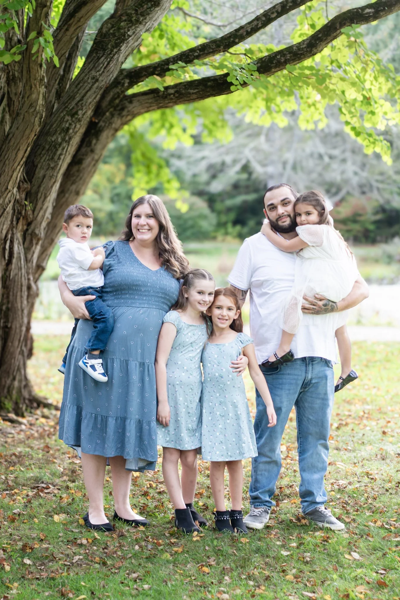 A family of six posing outdoors under a large tree with green leaves. The mother is wearing a blue dress and holding a young boy, five children are standing in front, and the father is holding a girl.