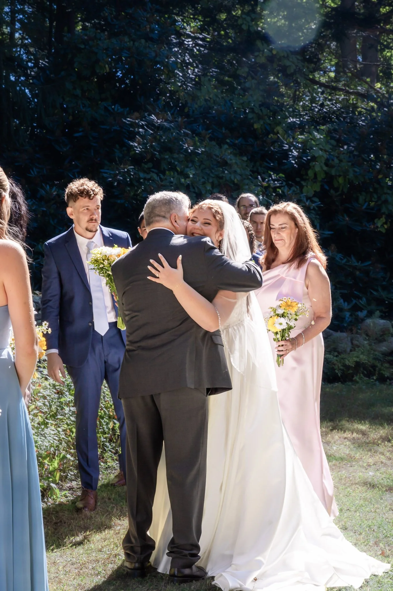 A bride and an older man hugging during an outdoor wedding ceremony. Bridesmaids and groomsmen are in the background.