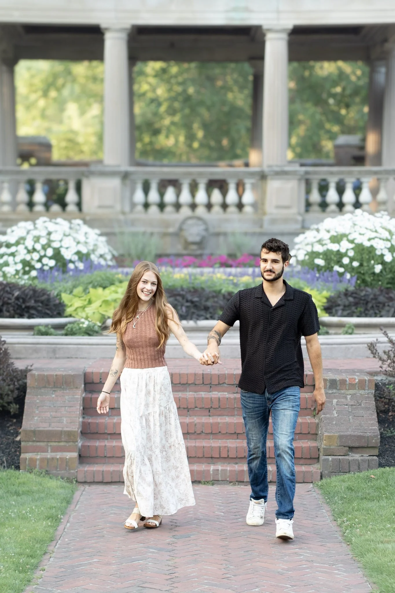 A young woman and man holding hands and walking down a brick pathway in front of a garden with flowers and a bridge in the background, in Beverly, MA