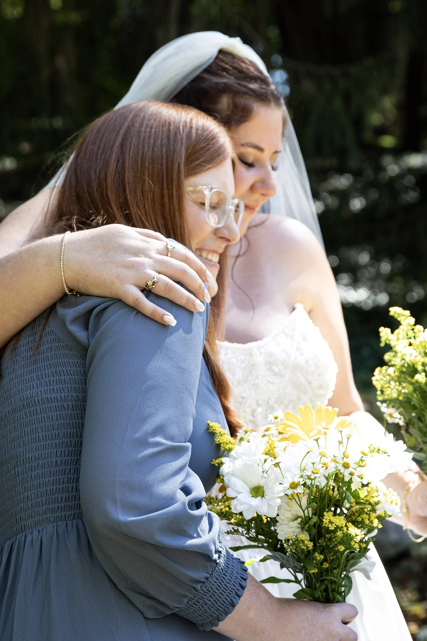 Two women, one in a wedding dress and veil, embracing and smiling, outdoors with greenery, one holding a bouquet of white and yellow flowers.