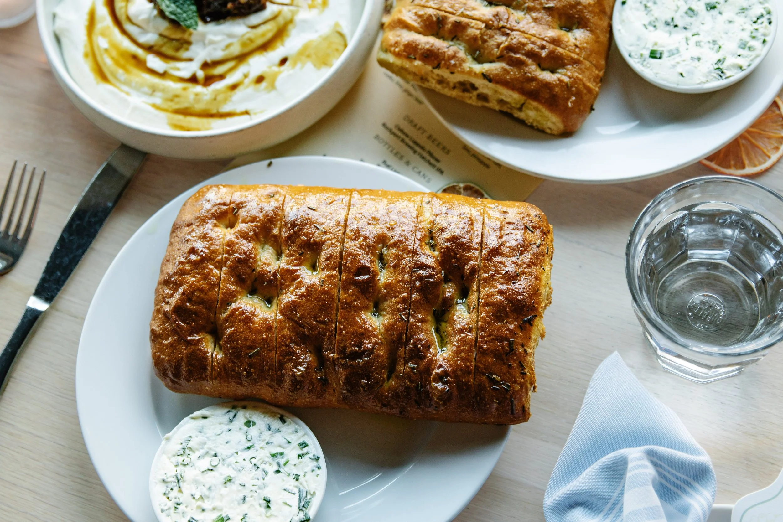 Close-up of a baked bread loaf on a white plate, accompanied by a small bowl of herb butter, a glass of water, and partially visible lemon slices, on a wooden table with a fork and knife to the left and a white napkin on the right.