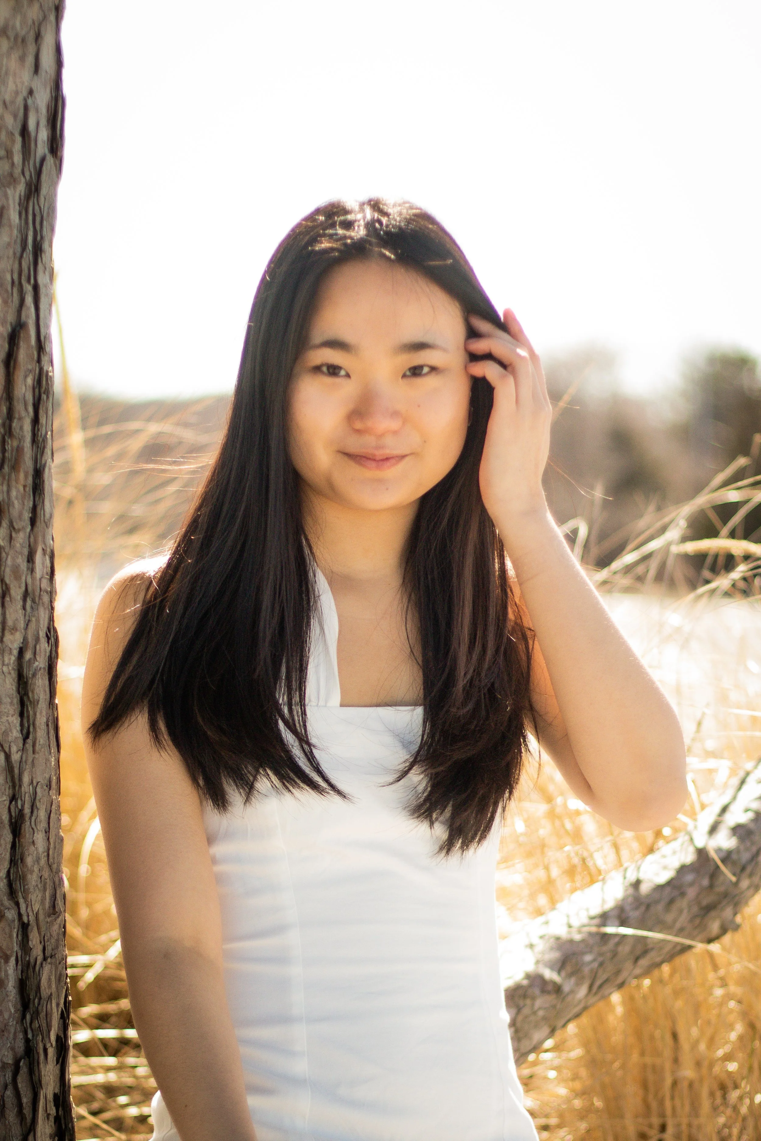 A young woman with long black hair standing outdoors near a tree in a sunlit field.