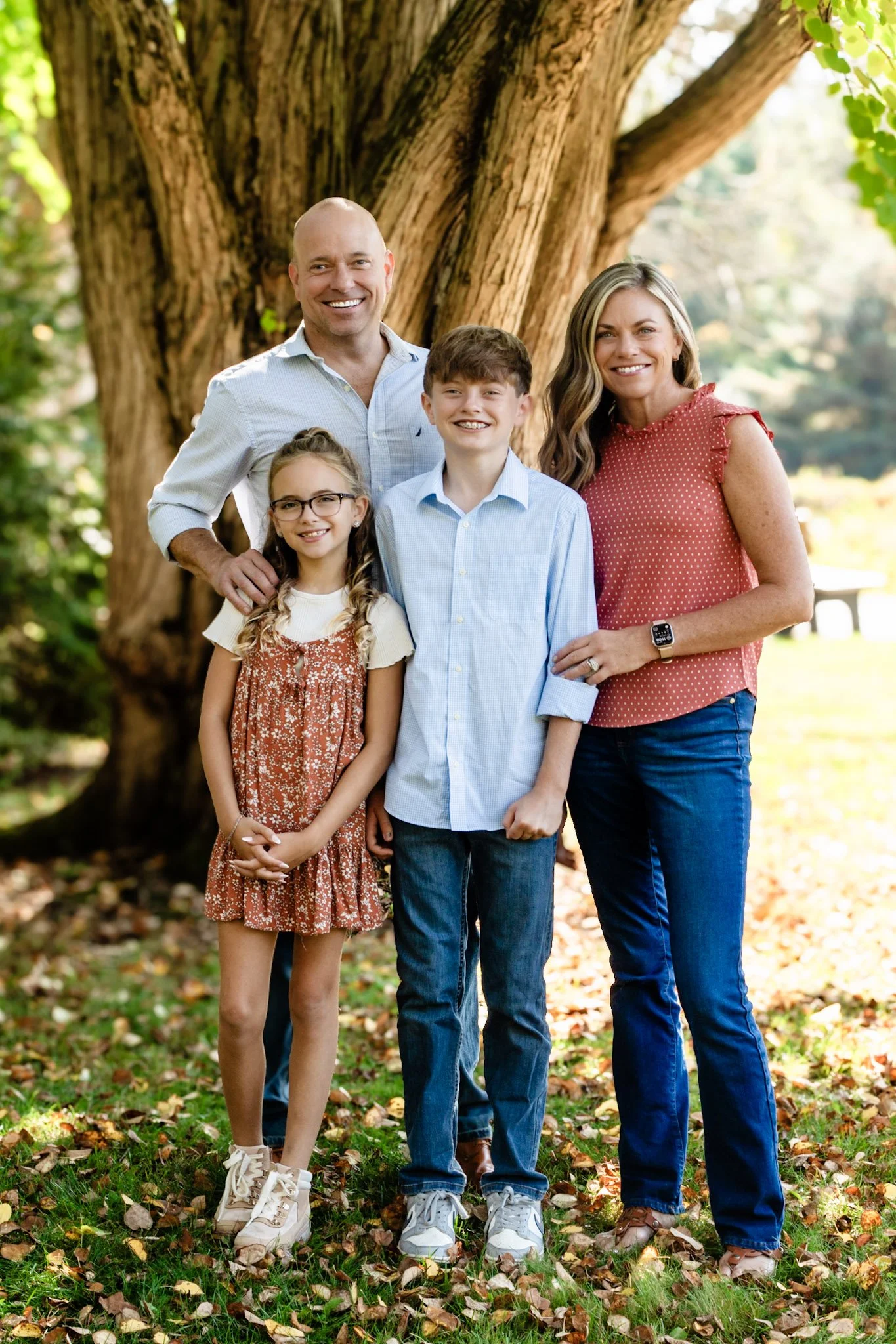 Benson Park, NH A family of five standing outdoors in a park with a large tree behind them, smiling, dressed casually, and surrounded by fallen autumn leaves.