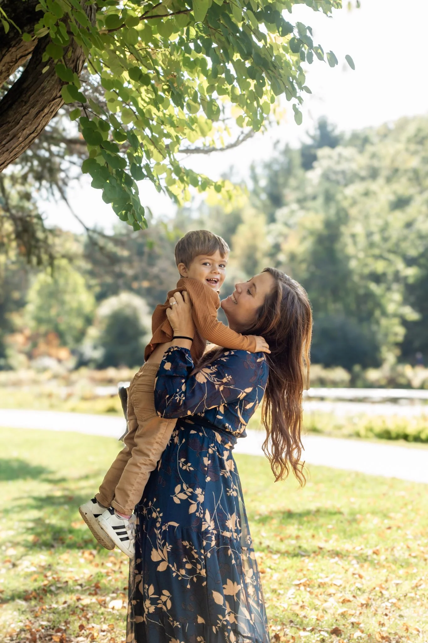 Hudson, NH, Mom and son photos. A woman lifting a young boy in the air outdoors in a park with trees and sunlight filtering through the leaves.
