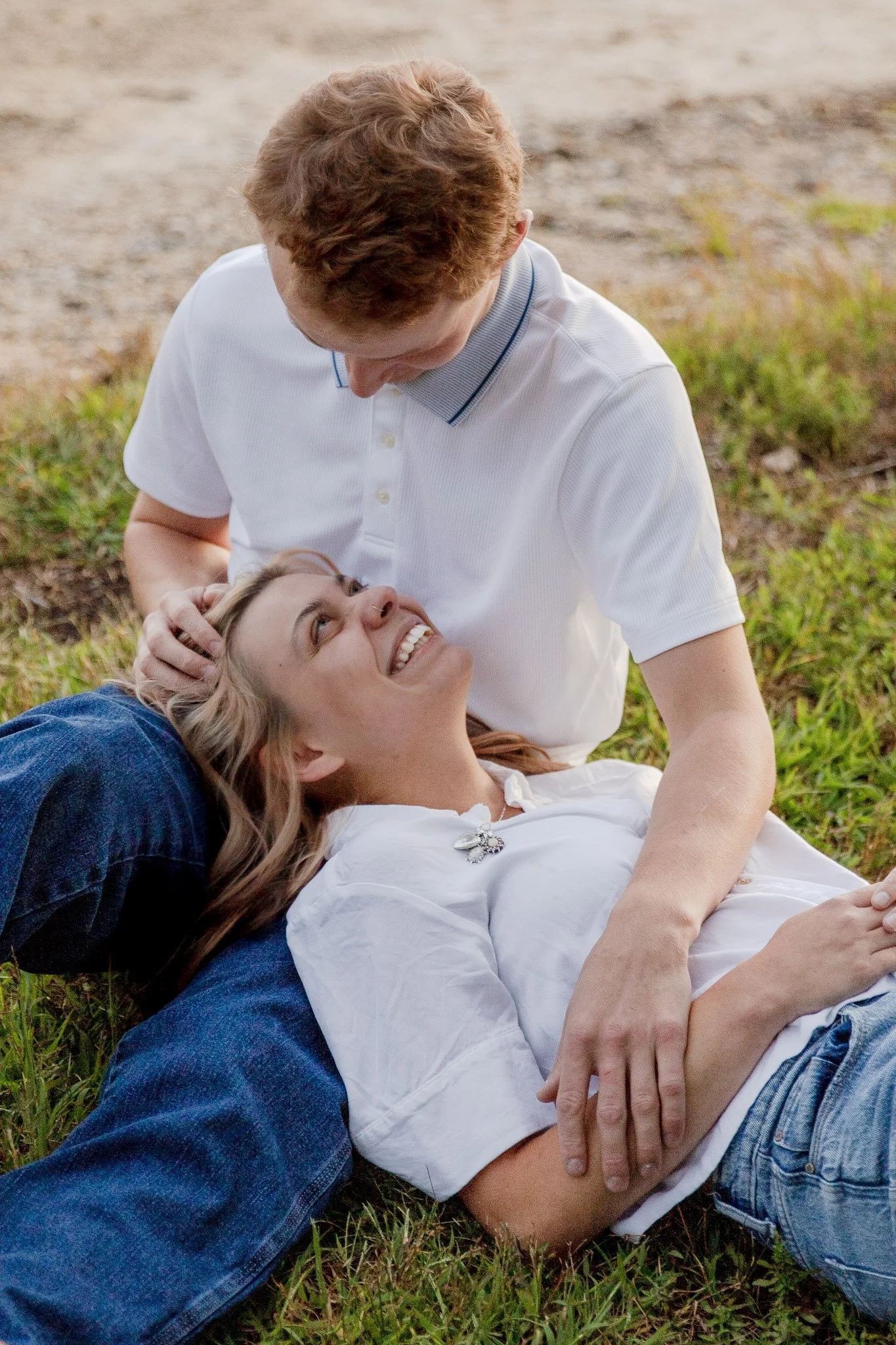 A man and woman are lying on the grass outdoors. The woman is smiling and looking up at the man, who is leaning over her. The scene is light and joyful.