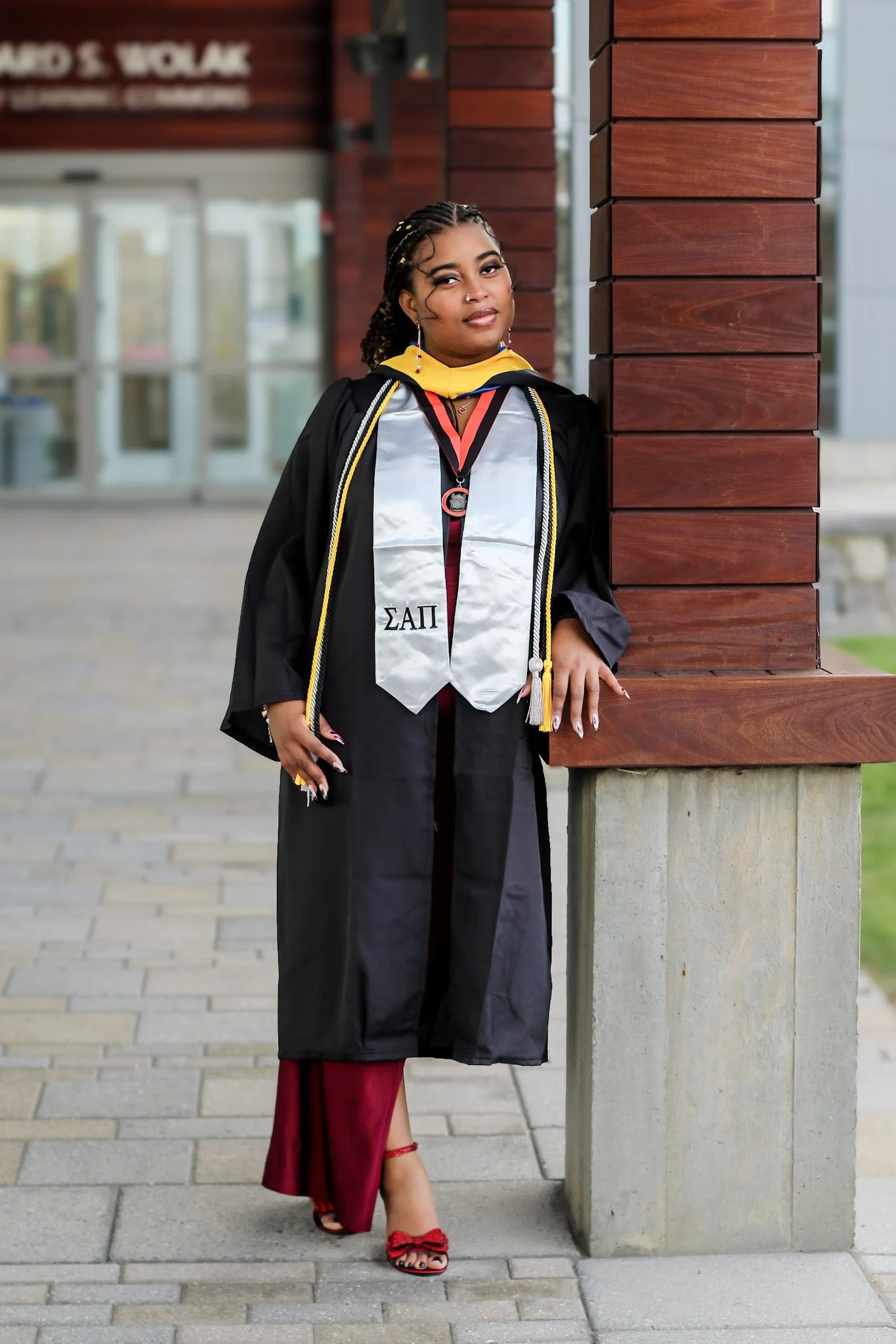 A woman in graduation regalia standing next to a wooden pillar outside during the day.