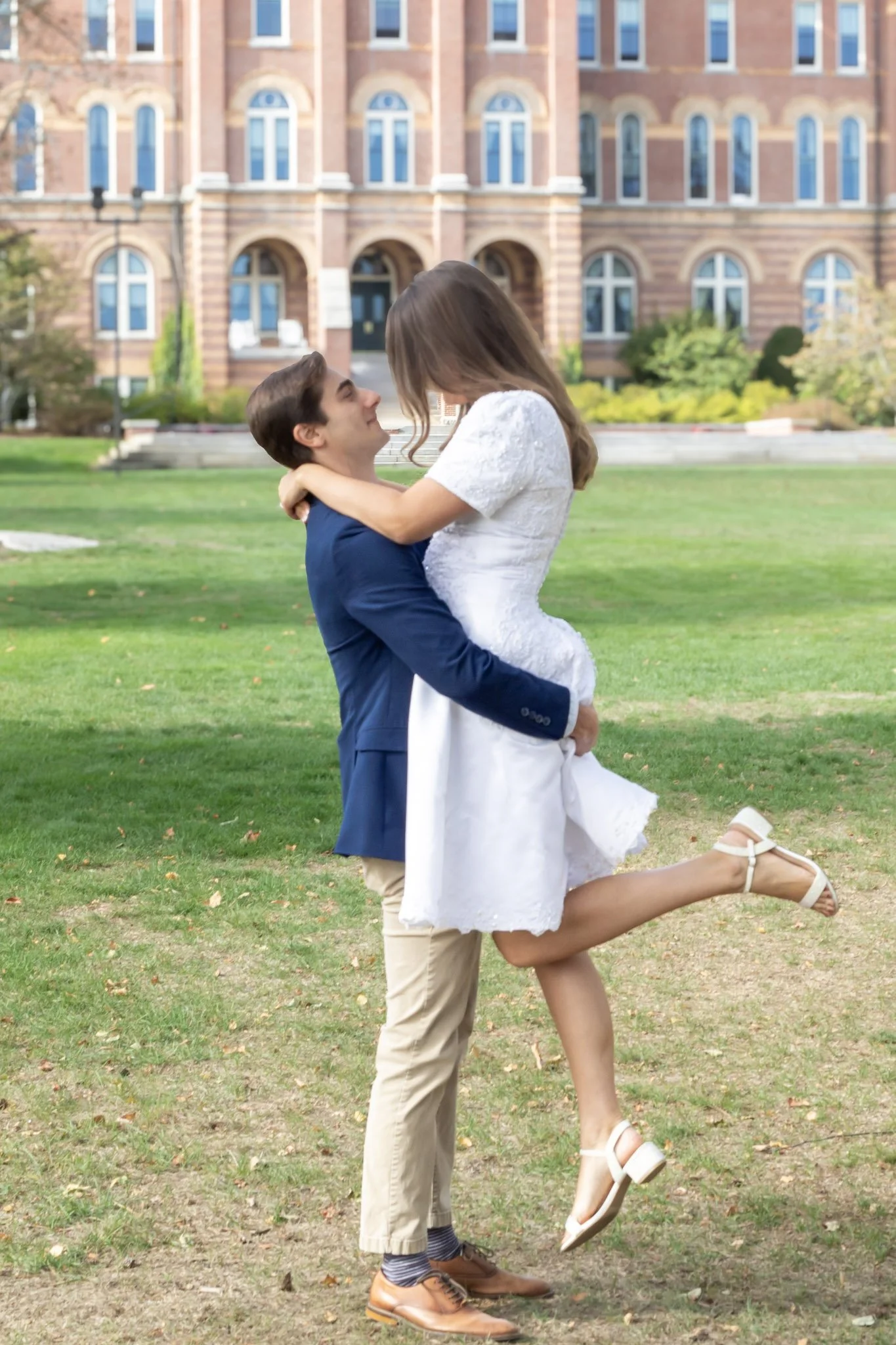 A man in a blue blazer lifting a woman in a white dress in a park with a large red brick building in the background.