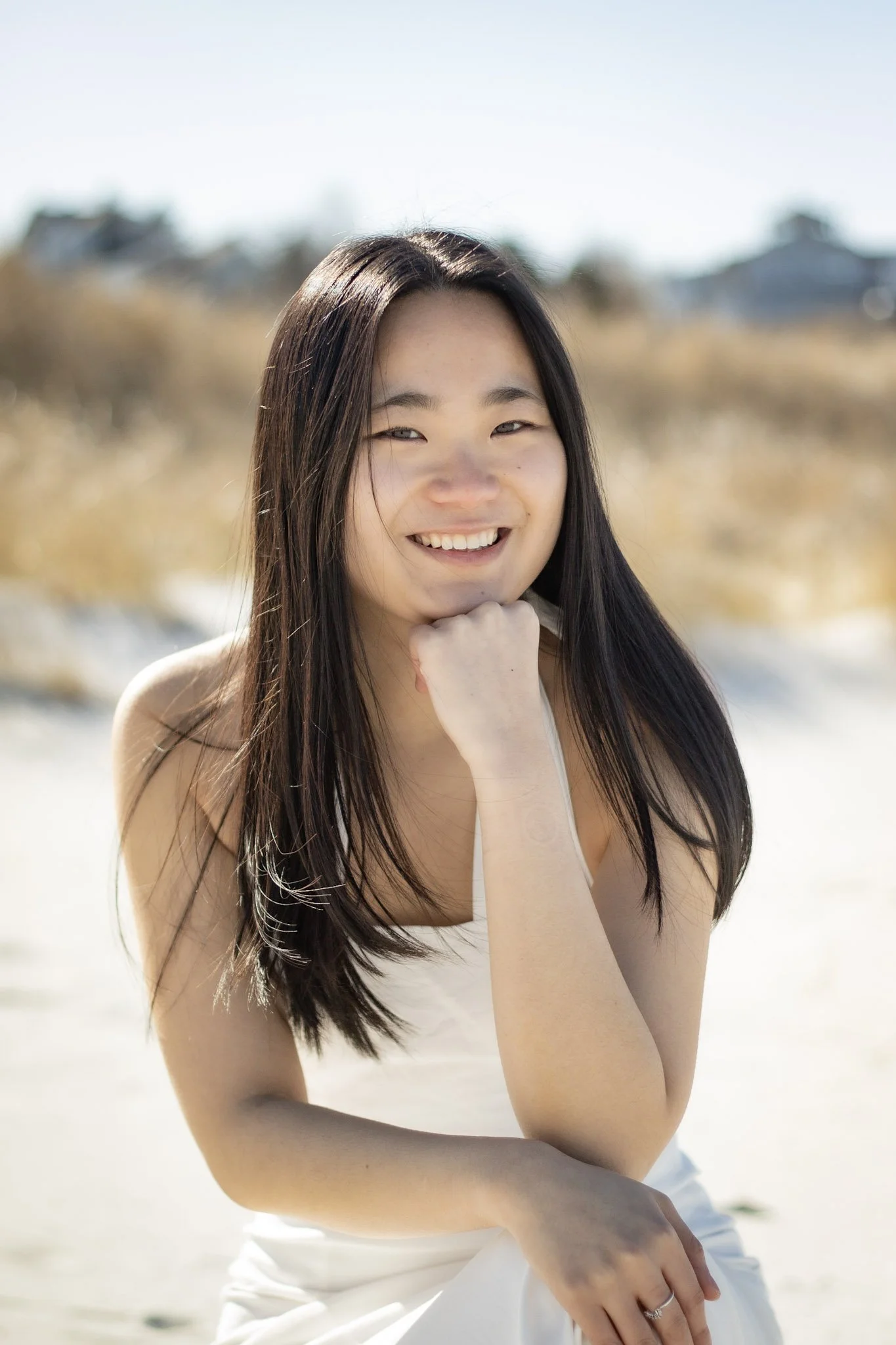 A young woman with long dark hair smiling and resting her chin on her hand outdoors on a sunny day.