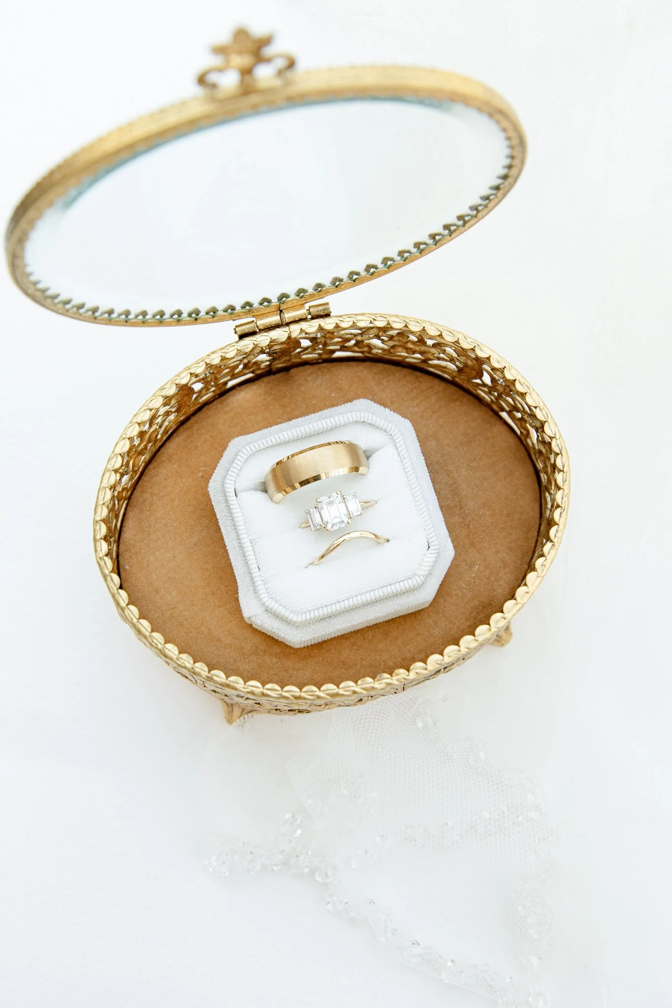 Cape Cod, MA Wedding Ring Photo, Open round gold jewelry box with a white velvet interior, containing a gold wedding band and a diamond engagement ring, resting on a white surface with lace fabric and scattered water crystal beads.