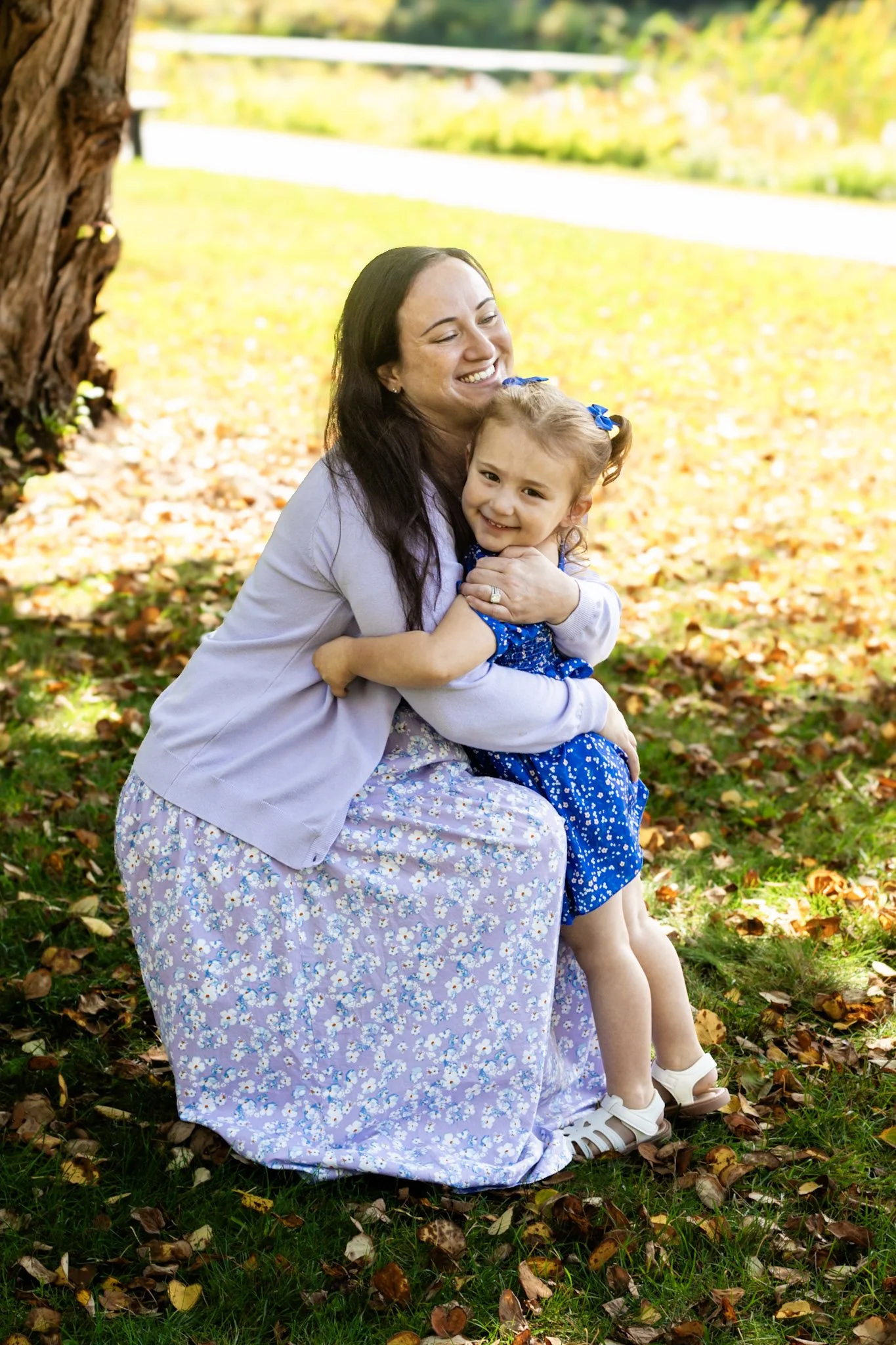 A woman and a girl are hugging outdoors on a sunny day, surrounded by fallen autumn leaves and a large tree.