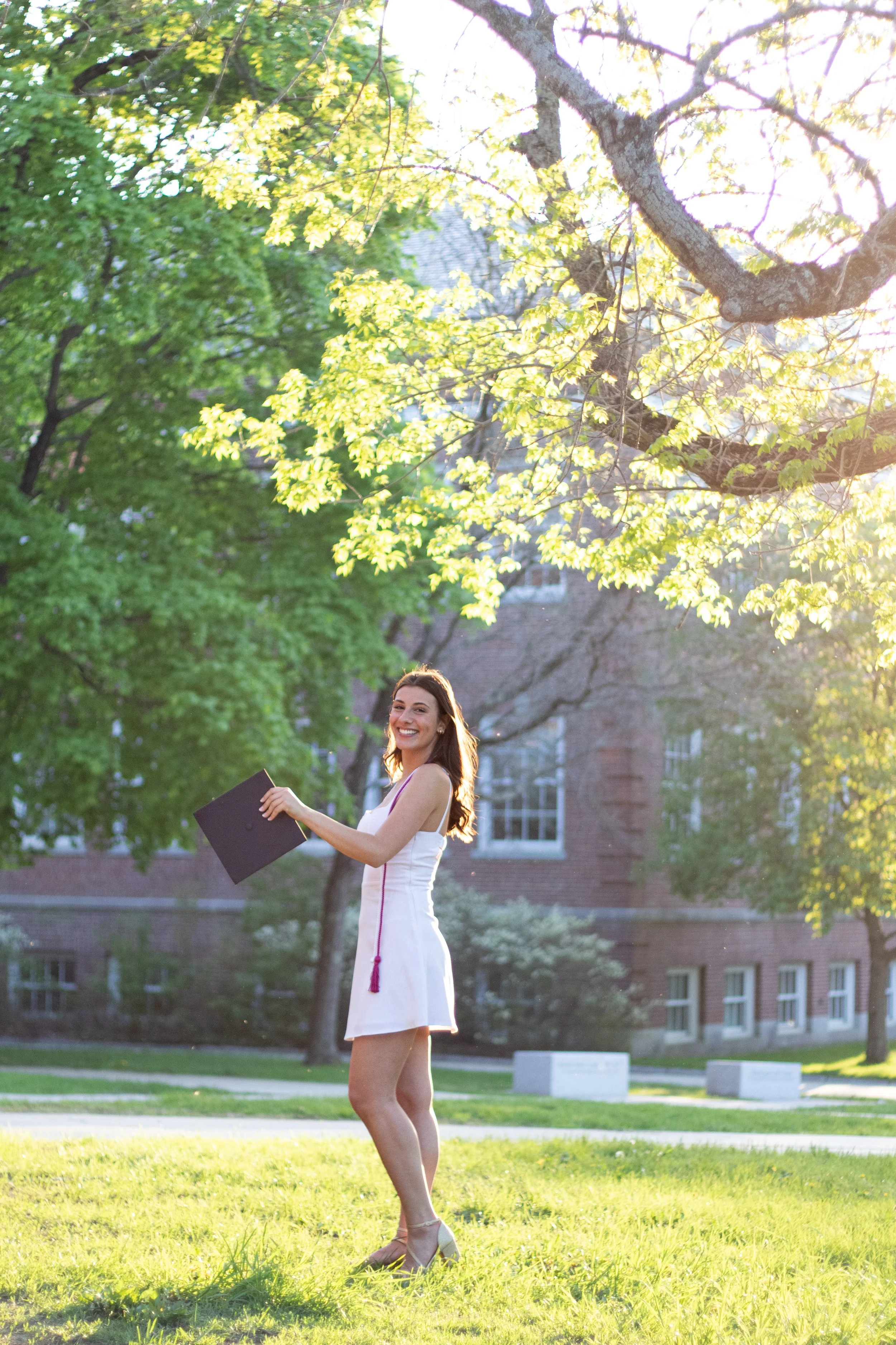 A young woman in a white dress holding a diploma standing on a grassy area outdoors, smiling and looking at the camera, with a college campus building in the background and sunlight filtering through trees.