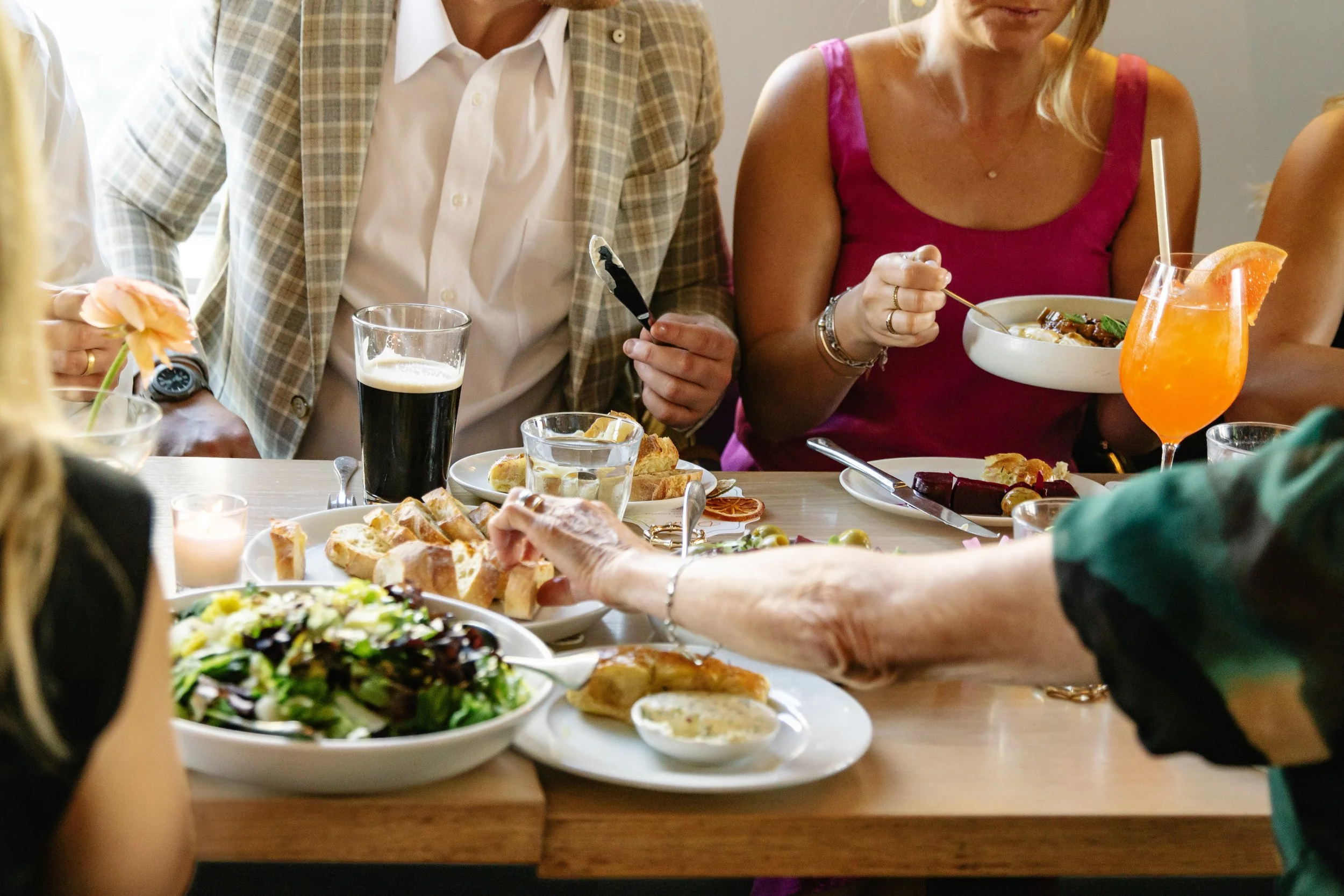 Boston, MA, Family and wedding guest eating tablescape image: People gathered around a dining table enjoying a meal with salads, bread, and drinks, including a dark beer and an orange cocktail, in a well-lit indoor setting.