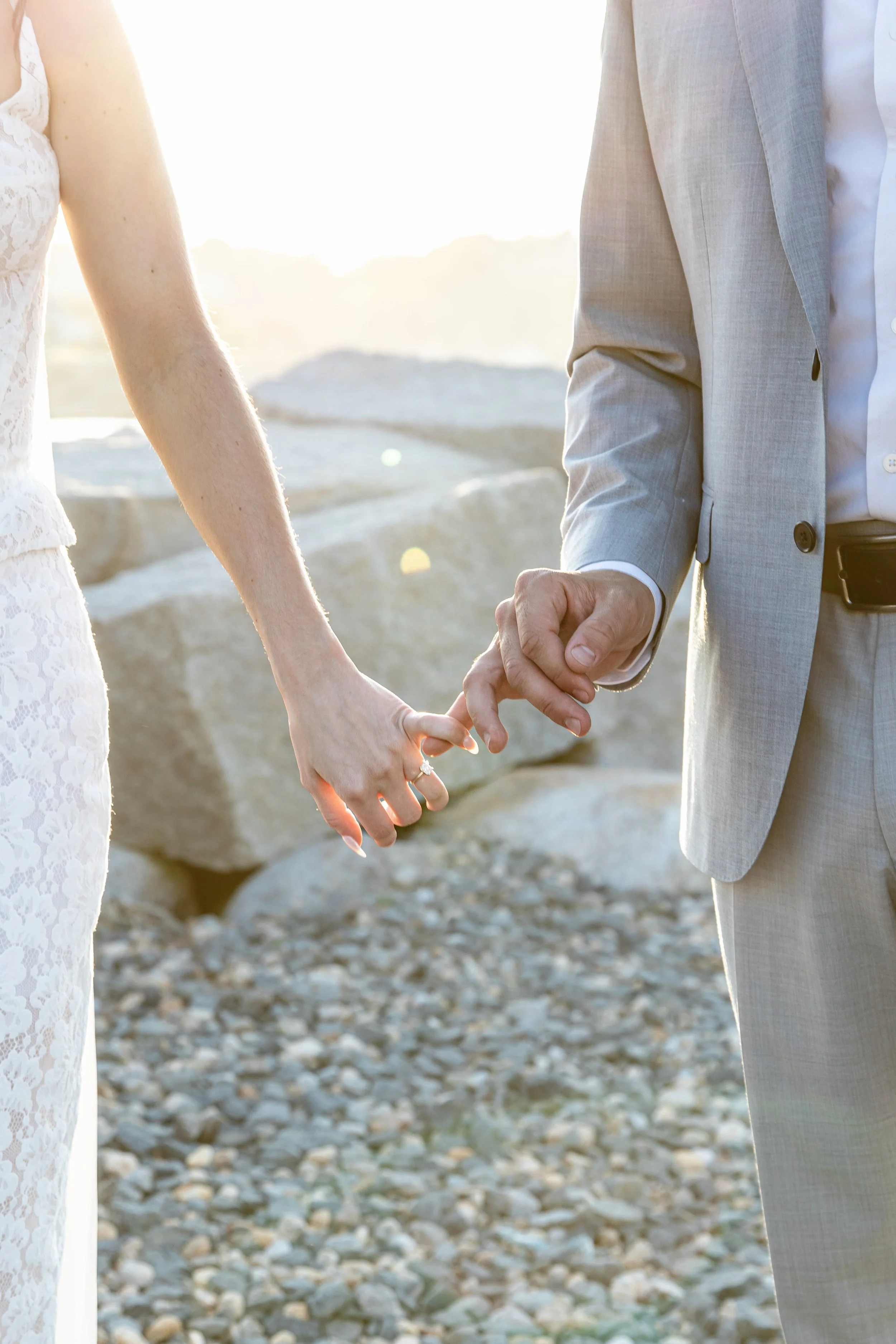 Close-up of a couple holding hands during their wedding, with a rocky beach and sunset in the background.
