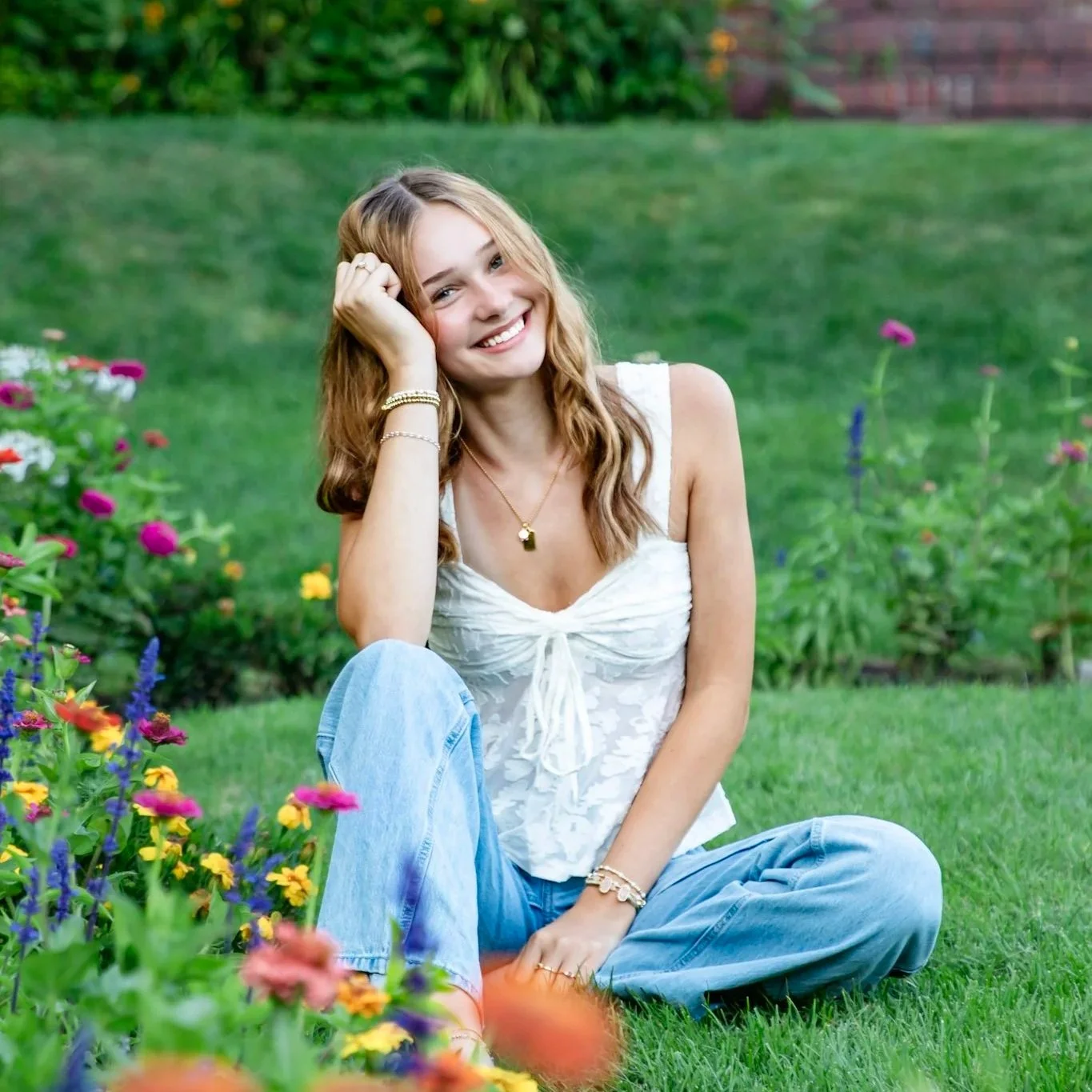 Boston, MA Senior Photos. A young woman with light brown hair and a bright smile sitting on the grass in a garden surrounded by colorful flowers, wearing a white sleeveless top and blue jeans.