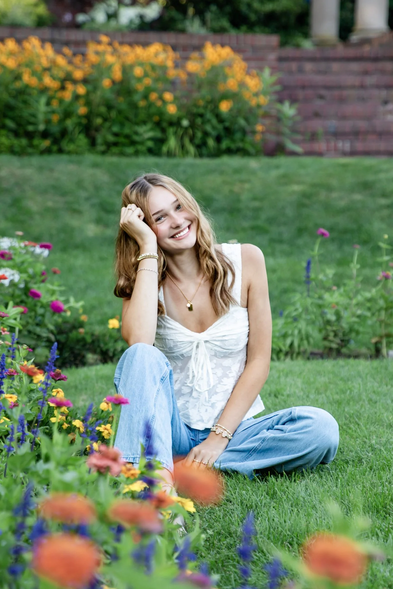 A smiling young woman with wavy blonde hair sitting on the grass among colorful flowers in a garden with a brick wall and greenery in the background.