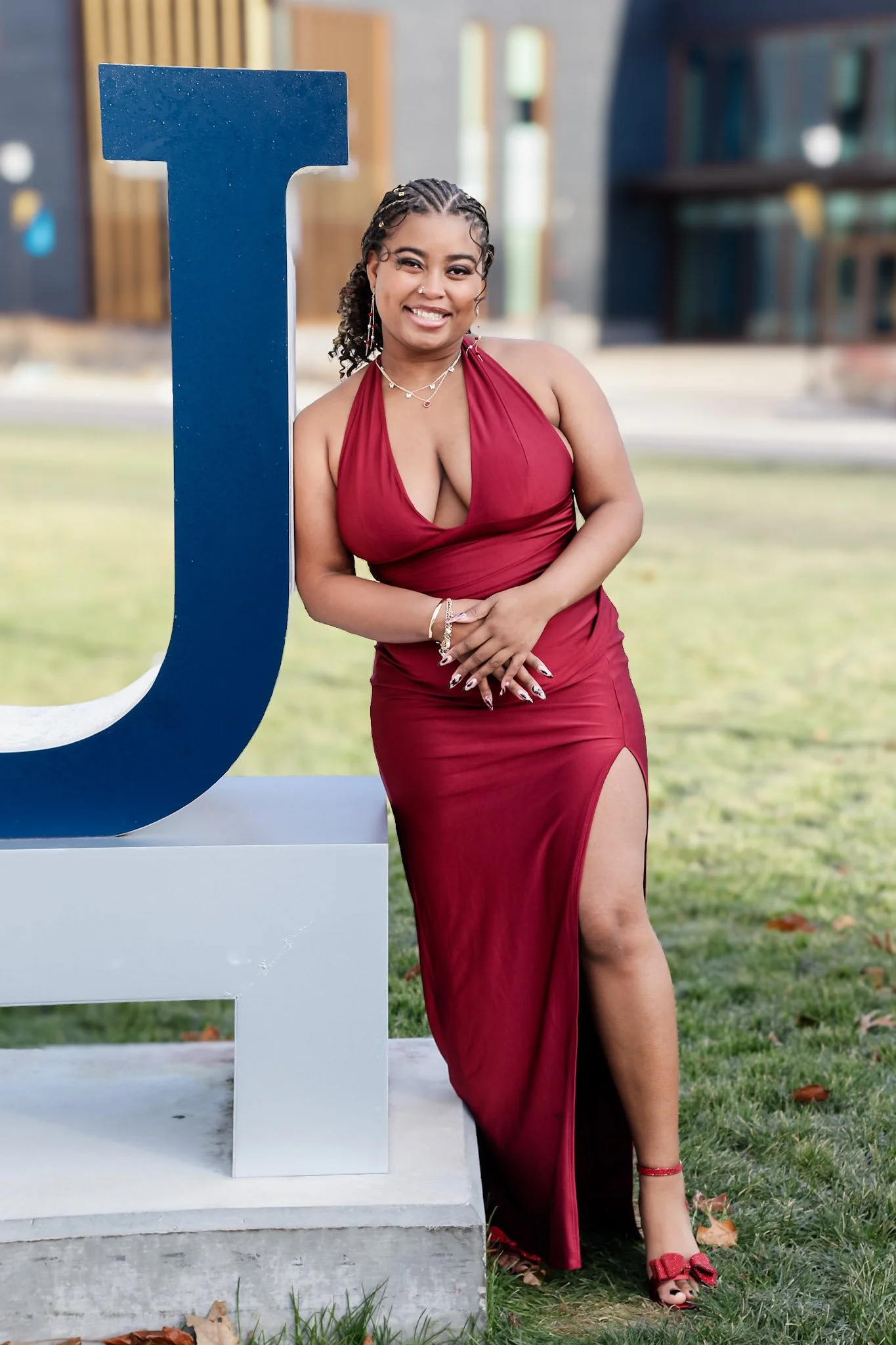 A woman in a red evening gown with a thigh-high slit and matching red heels, standing next to a large blue letter 'J' on a pedestal outdoors, smiling at the camera.