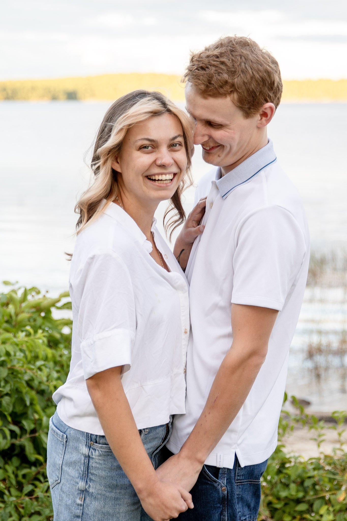 A happy couple standing outdoors by the water, holding hands, and smiling at each other.