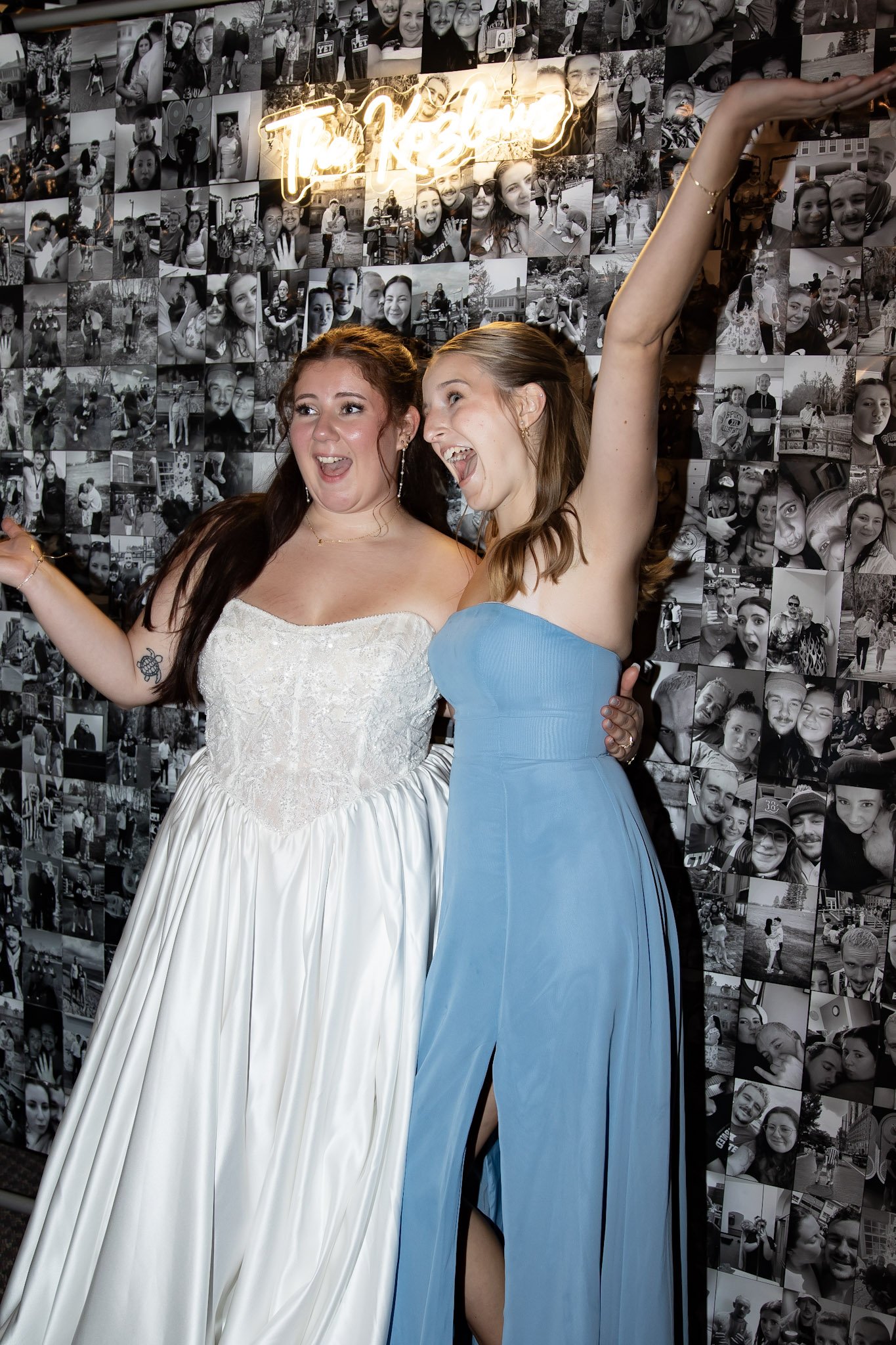 Two women celebrating at a party, one in a white dress and the other in a blue dress, smiling and posing with their arms raised, in front of a wall covered with photos and a neon sign.